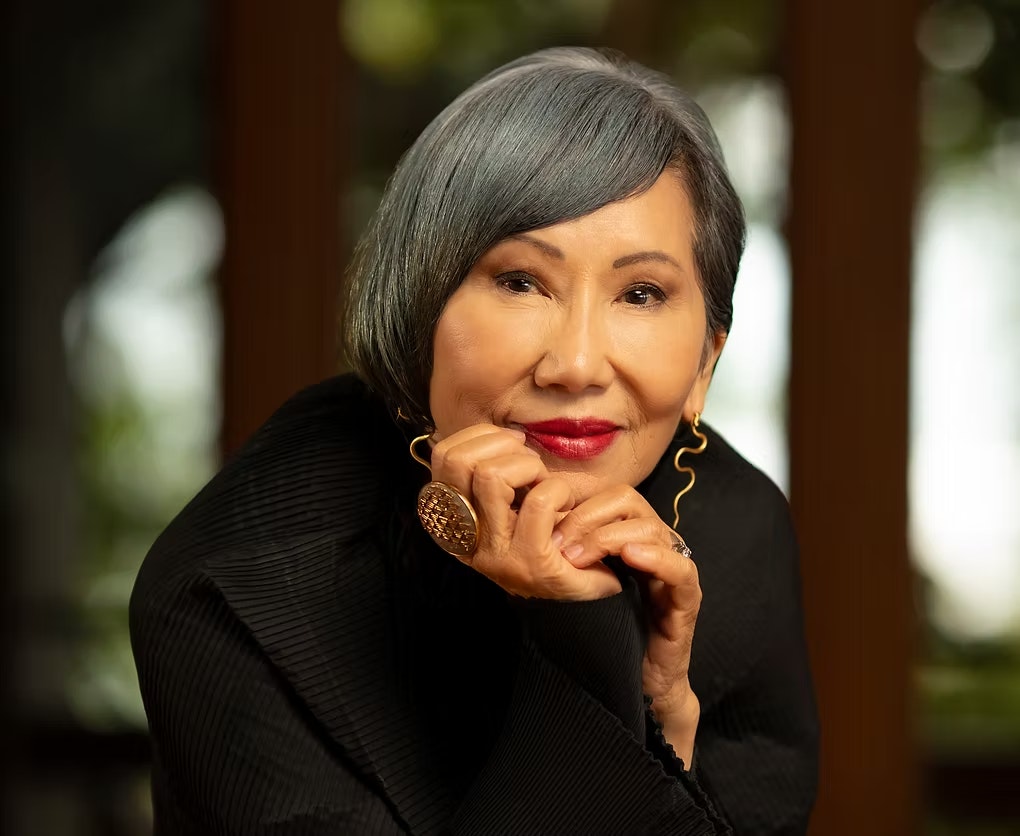 A person with short gray hair, wearing a black top and gold earrings, rests their chin on their hands, looking directly at the camera.