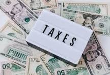 The image shows a sign saying "TAXES" placed on top of scattered U.S. dollar bills.