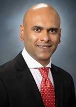 The image shows a professionally dressed man with a bald head, wearing a suit and tie, posing for a headshot.
