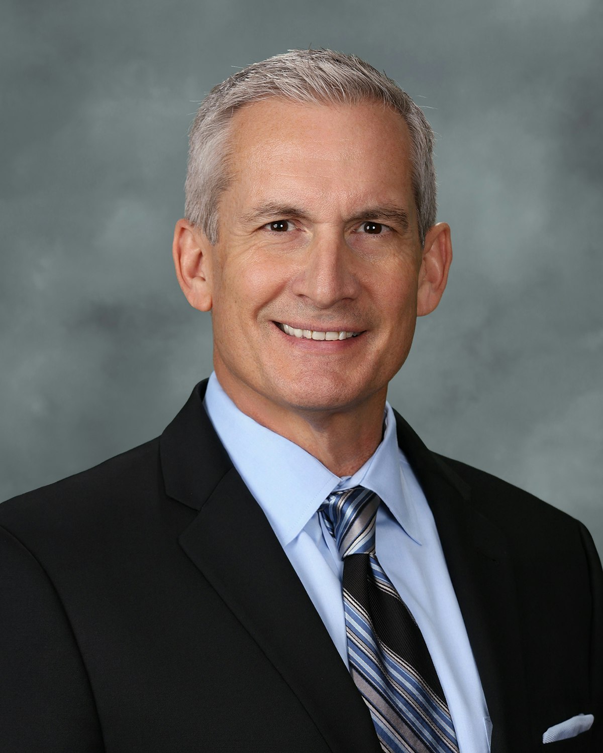 A professional headshot of an older man in a suit with a light blue shirt and striped tie, smiling against a gray background.