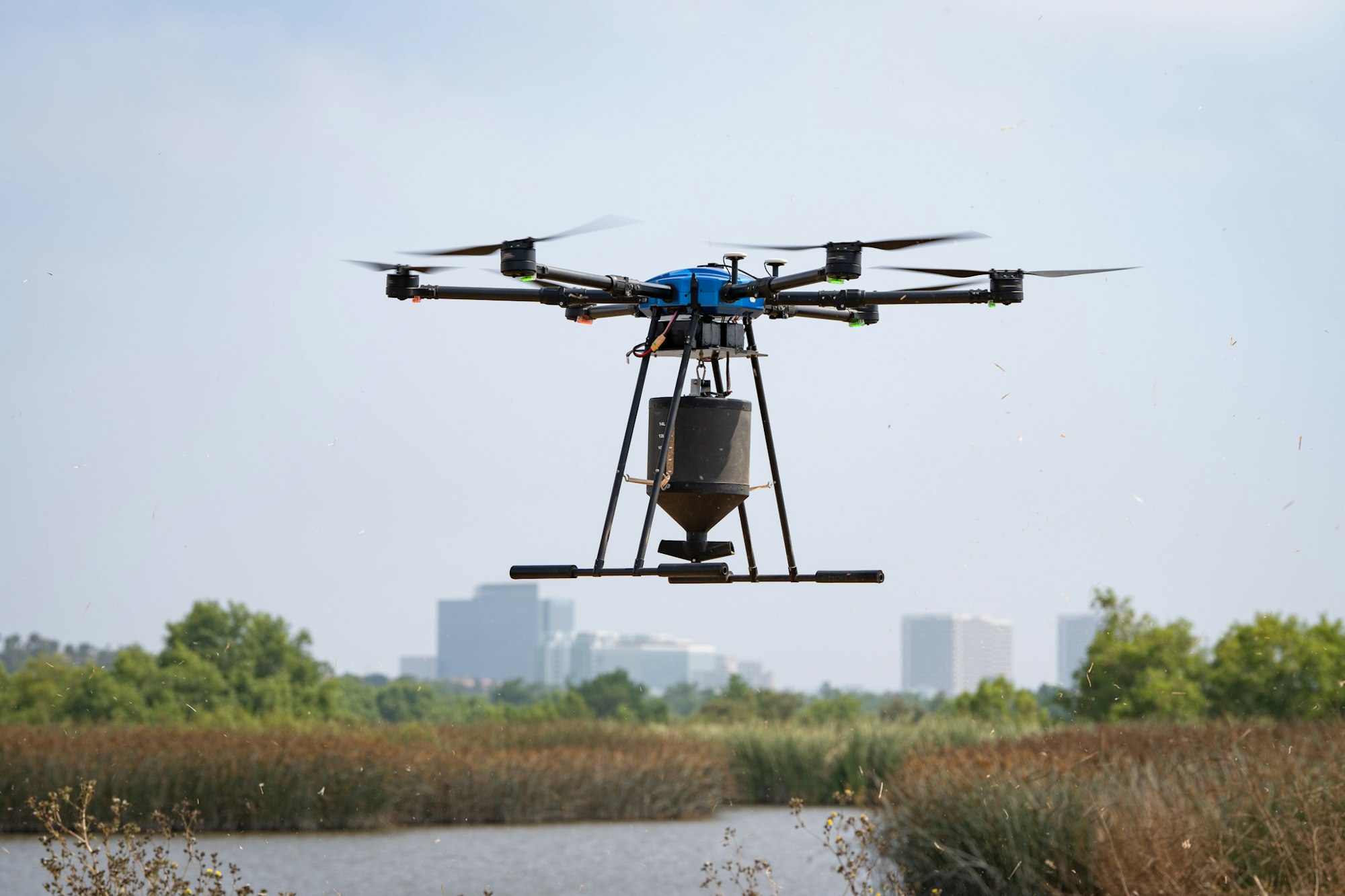 A drone with a payload, flying over a natural landscape with water and vegetation, city skyline in the background.