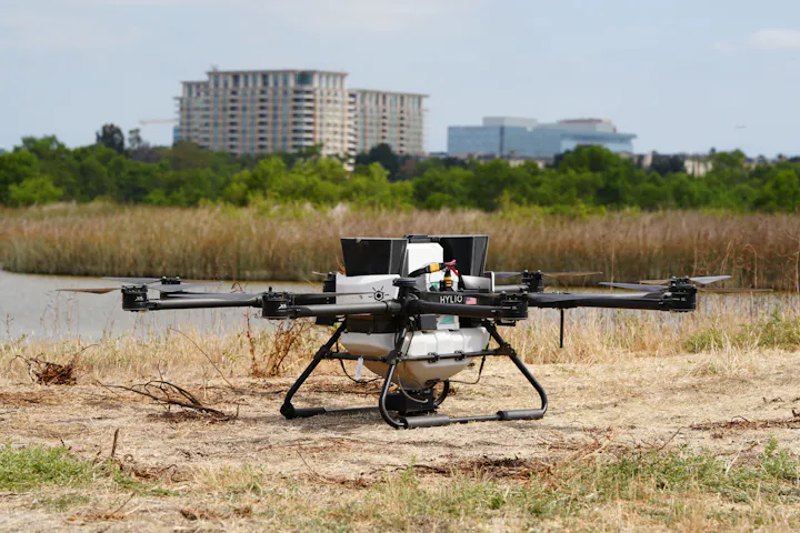 A drone with multiple rotors is positioned on the ground near a body of water and greenery, with buildings visible in the background.