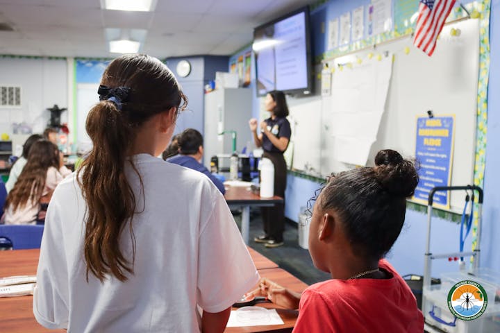 Classroom with a woman speaking to students and two girls are in the foreground.