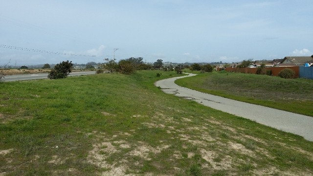 A winding pathway through grassy terrain, cloudy sky, some buildings in the distance.