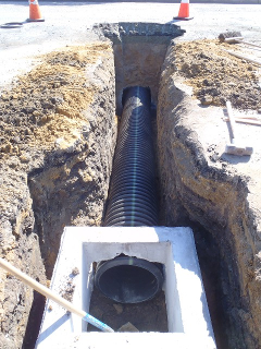 A trench with a large pipe and a concrete structure, possibly for utilities or drainage, with safety cones around the site.