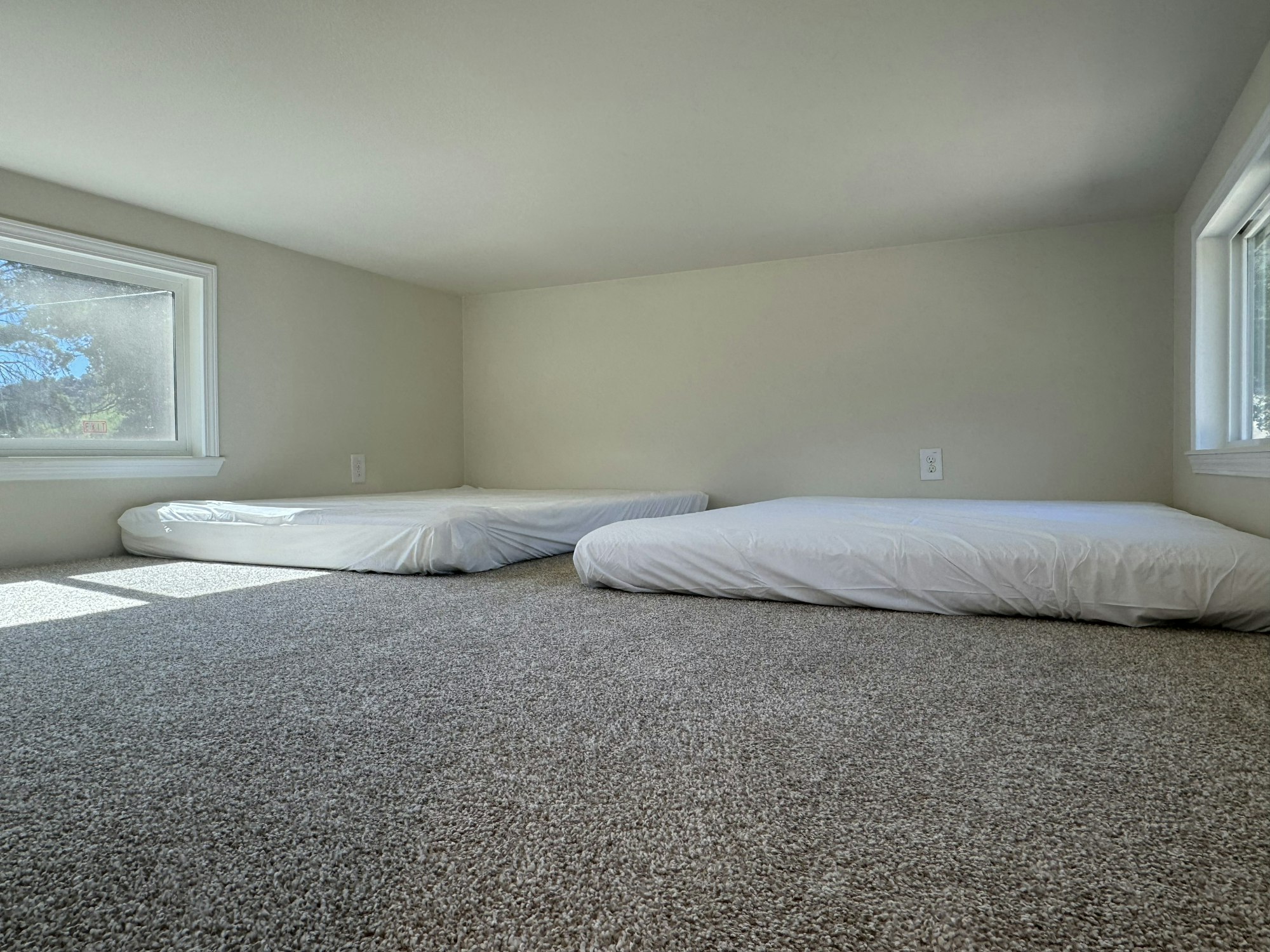 A small room with two low mattresses on a carpeted floor, near two windows.