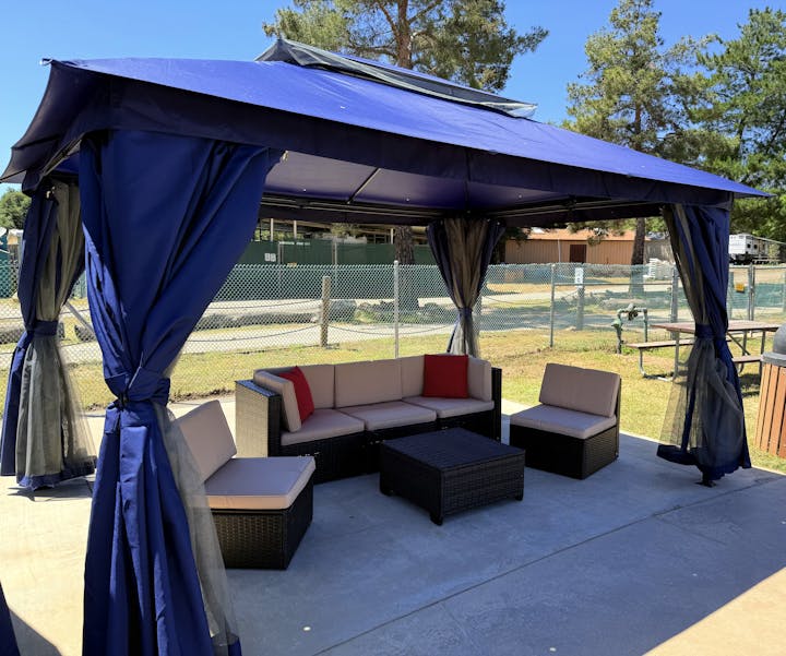 A gazebo with blue curtains, featuring outdoor seating including a corner sofa, chairs, and a table, set in a sunny garden area.