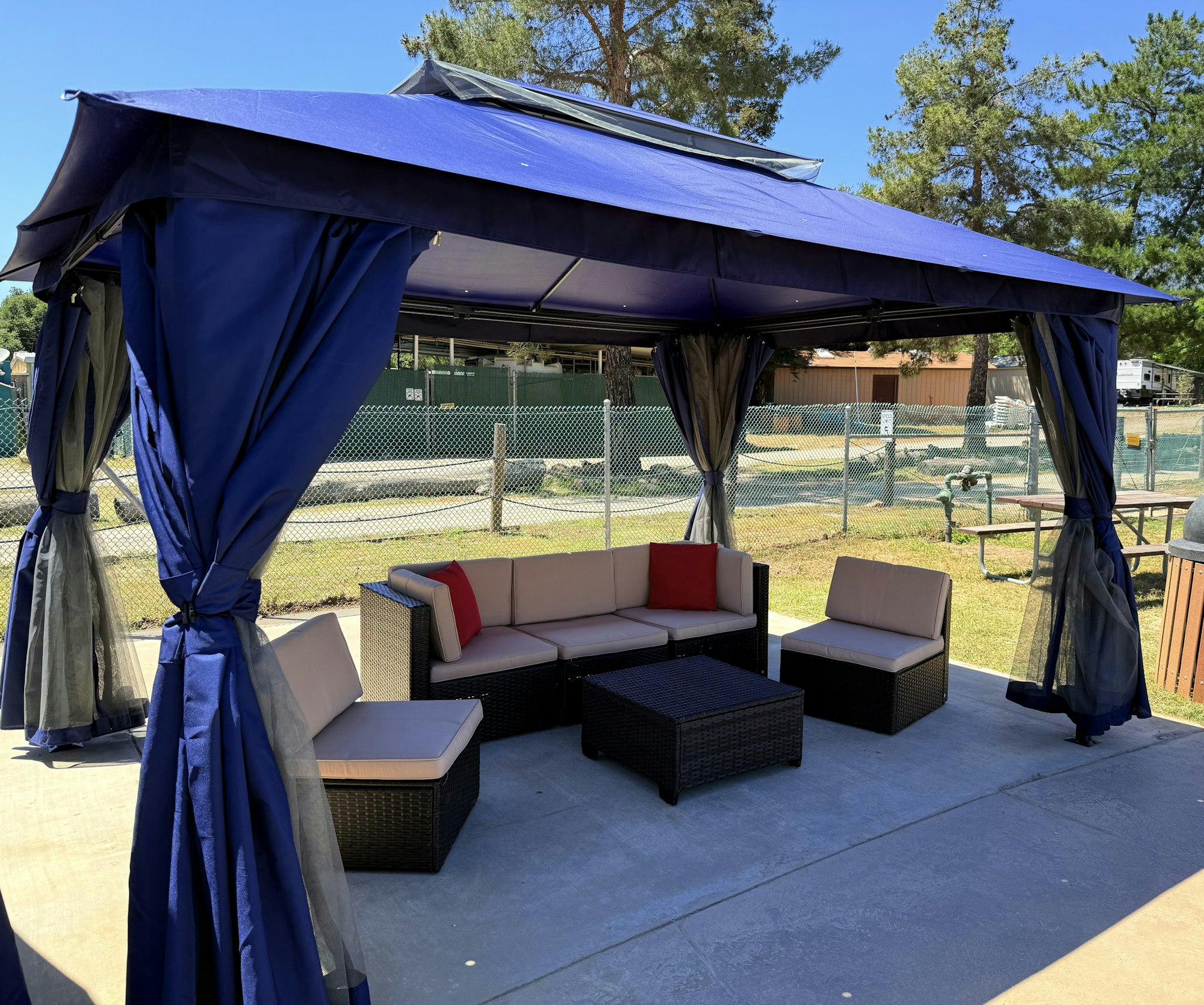 A gazebo with blue curtains, featuring outdoor seating including a corner sofa, chairs, and a table, set in a sunny garden area.