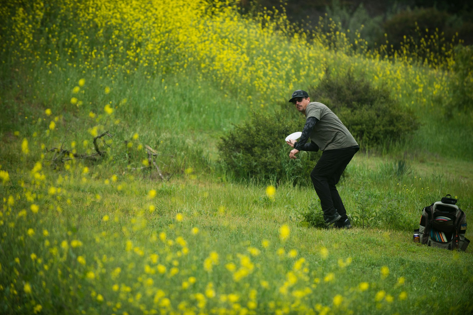 A person playing disc golf in a grassy field with yellow flowers, holding a disc and a backpack nearby.