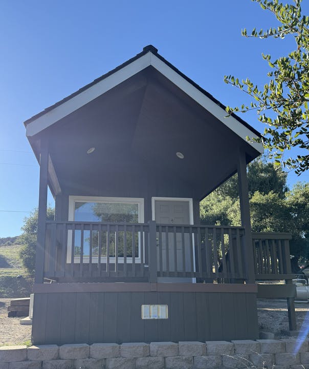 A small, elevated cabin with a porch and railing, set in a natural, sunny environment with trees.