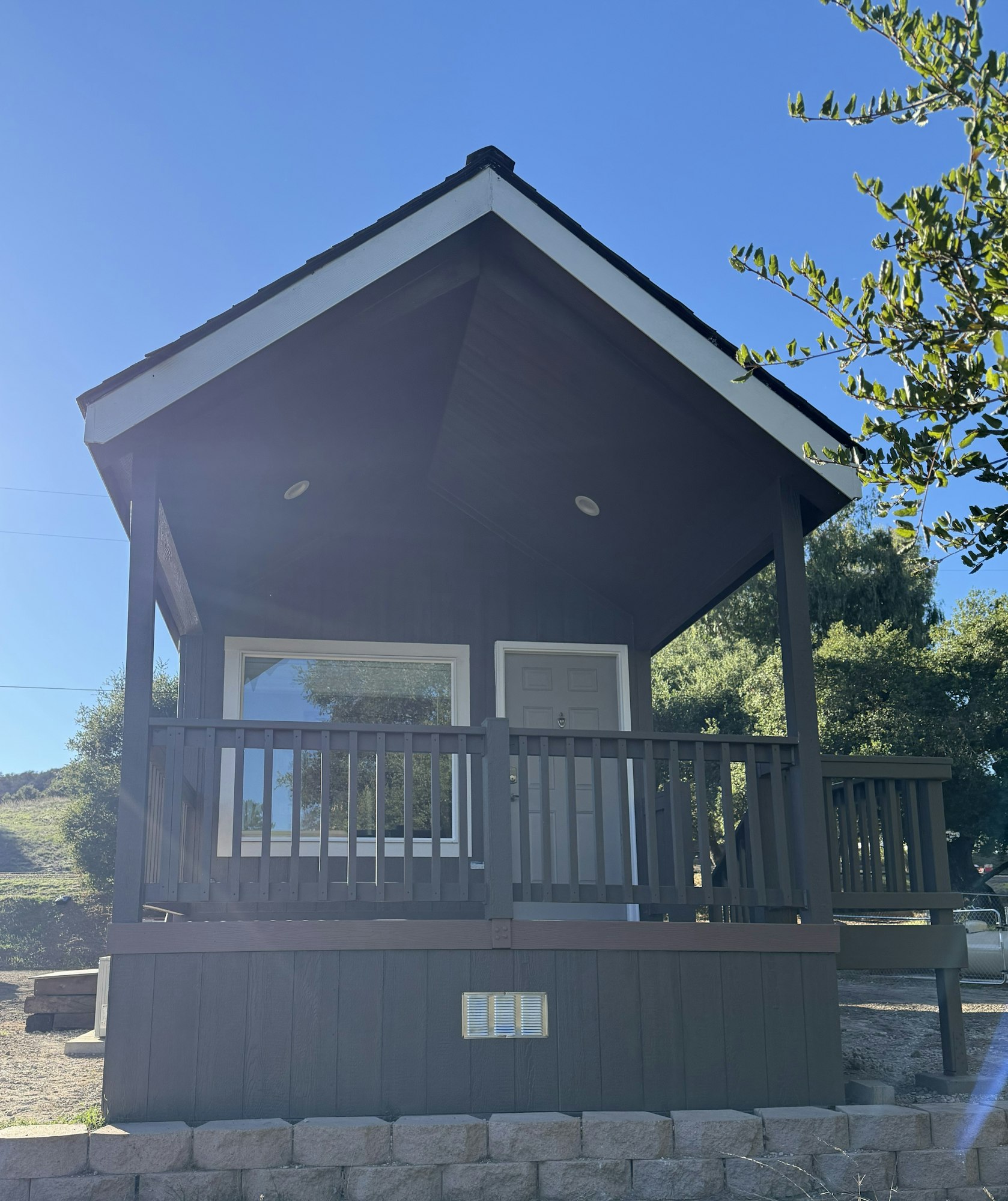 A small, elevated cabin with a porch and railing, set in a natural, sunny environment with trees.
