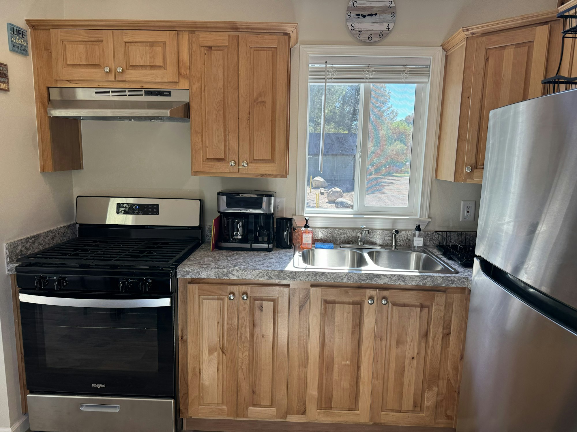 A kitchen with a stove, stainless steel fridge, wooden cabinets, coffee maker, sink, and a window with a view outside.