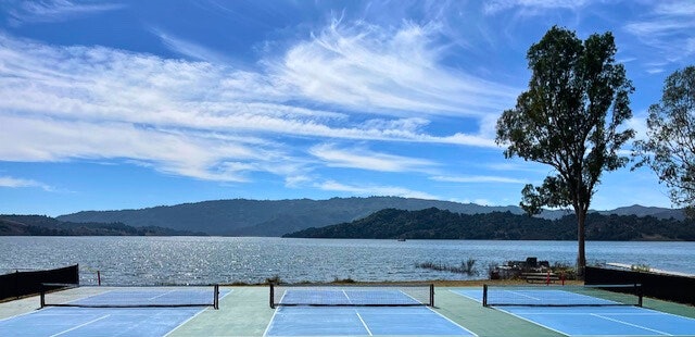 A scenic outdoor spot with tennis courts overlooking a lake and mountains under a bright sky with streaky clouds.