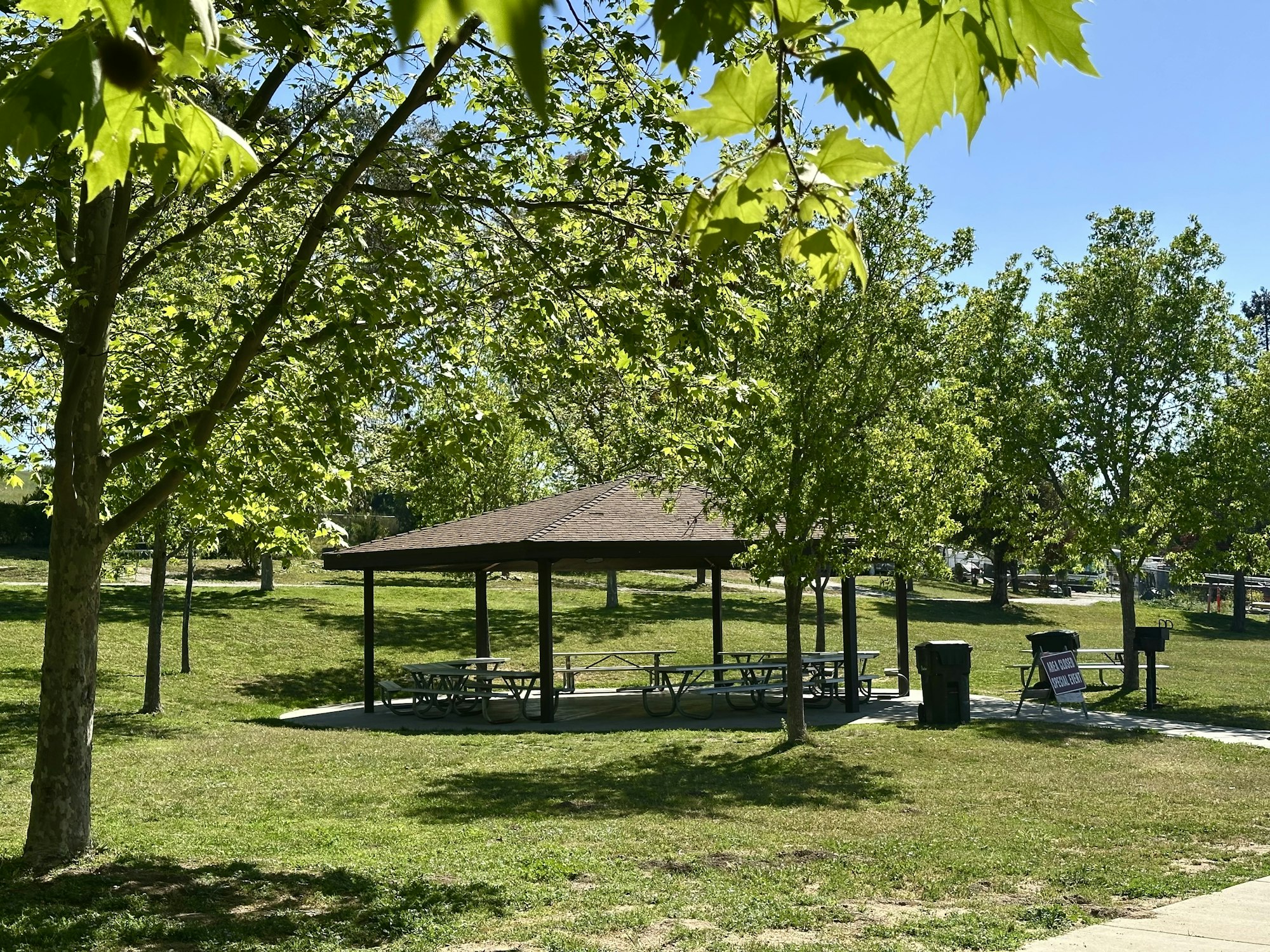 A park with trees, a wooden shelter, picnic tables, trash bins, and a sign on a grassy area.