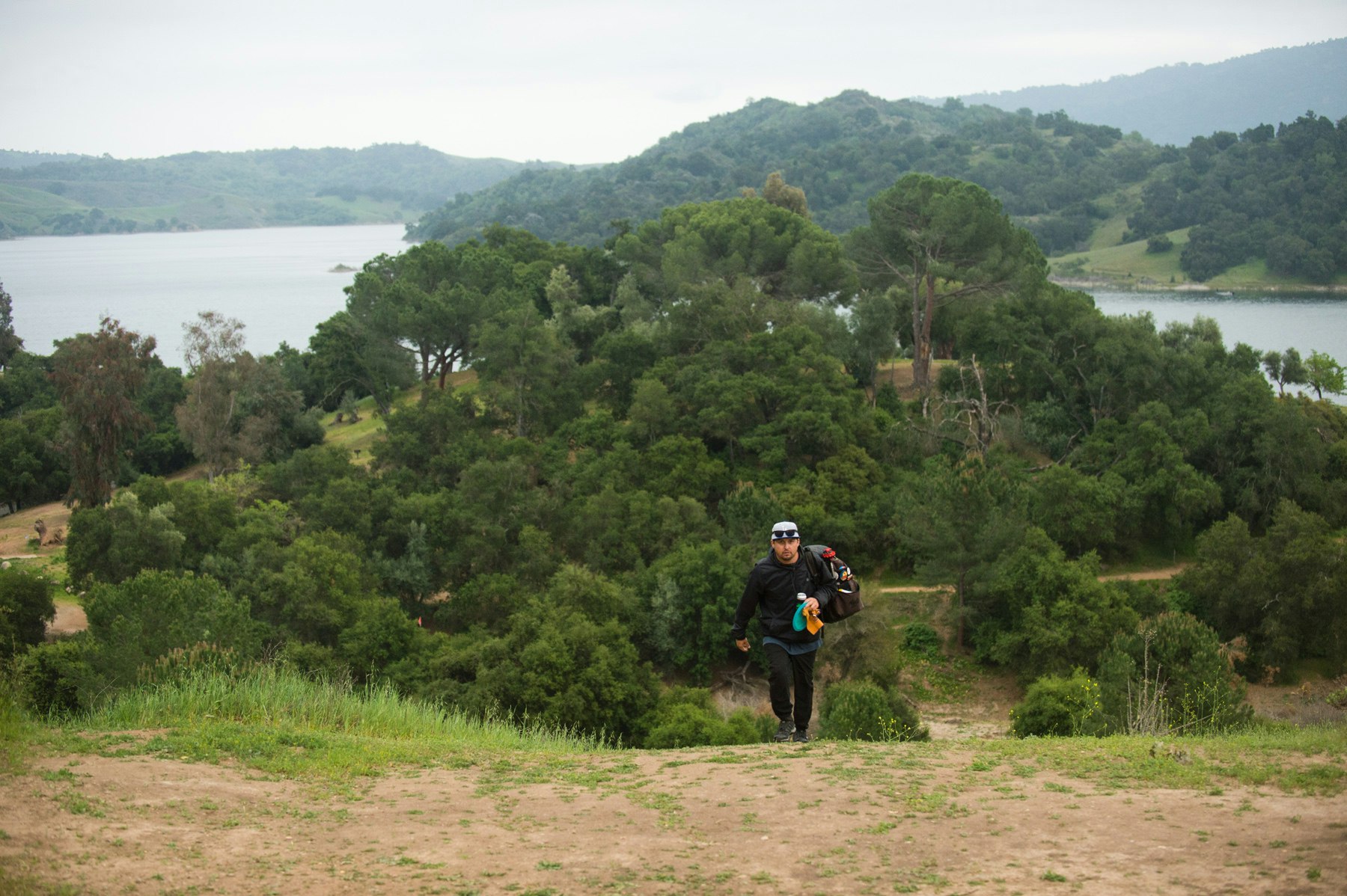 Person hiking uphill with a backpack, surrounded by lush greenery and a lake in the background.