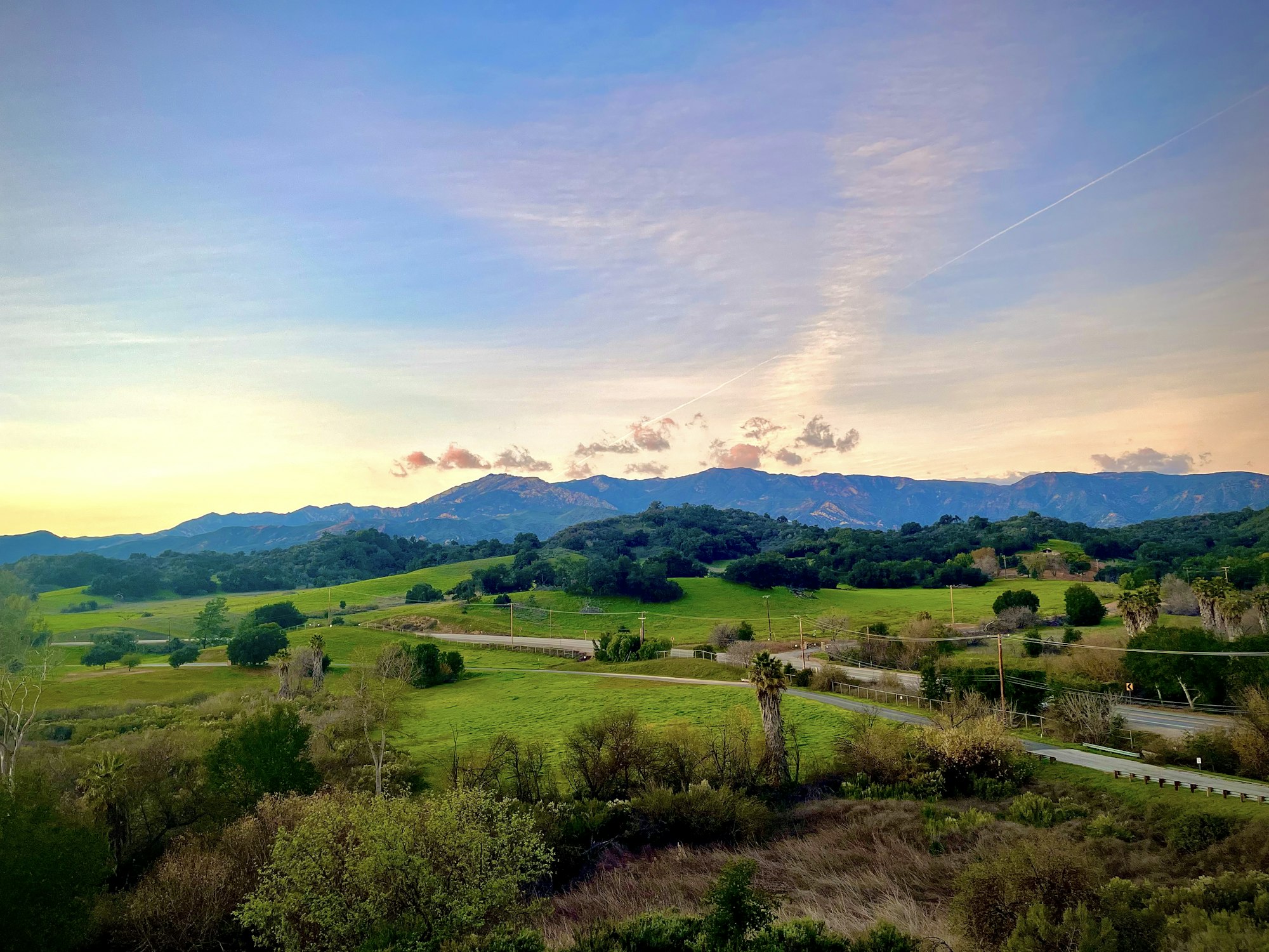 Scenic landscape with green fields, trees, and mountains under a colorful sky at sunset.