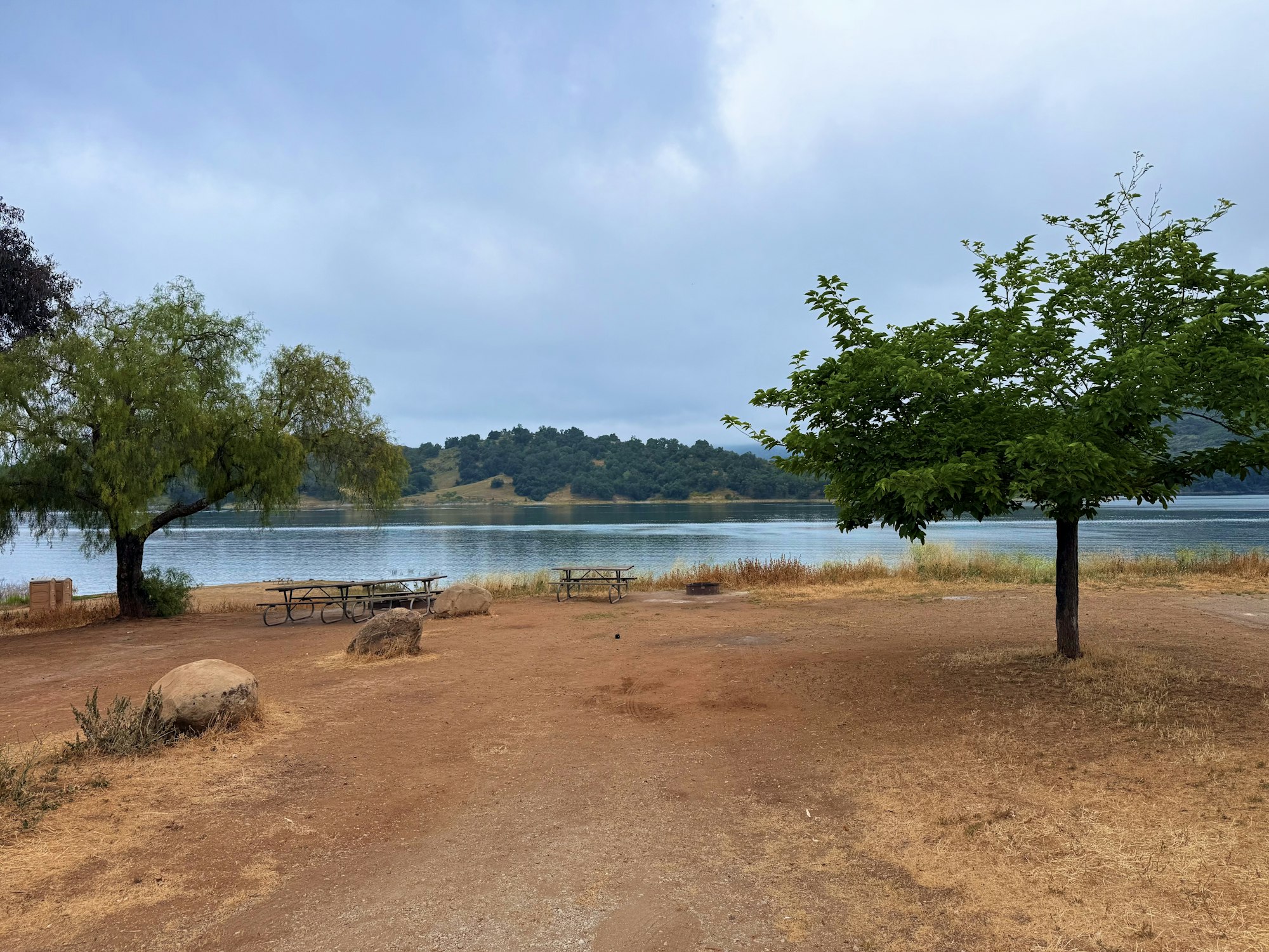 A scenic lakeside view with picnic tables, trees, and a grassy shoreline under a cloudy sky.