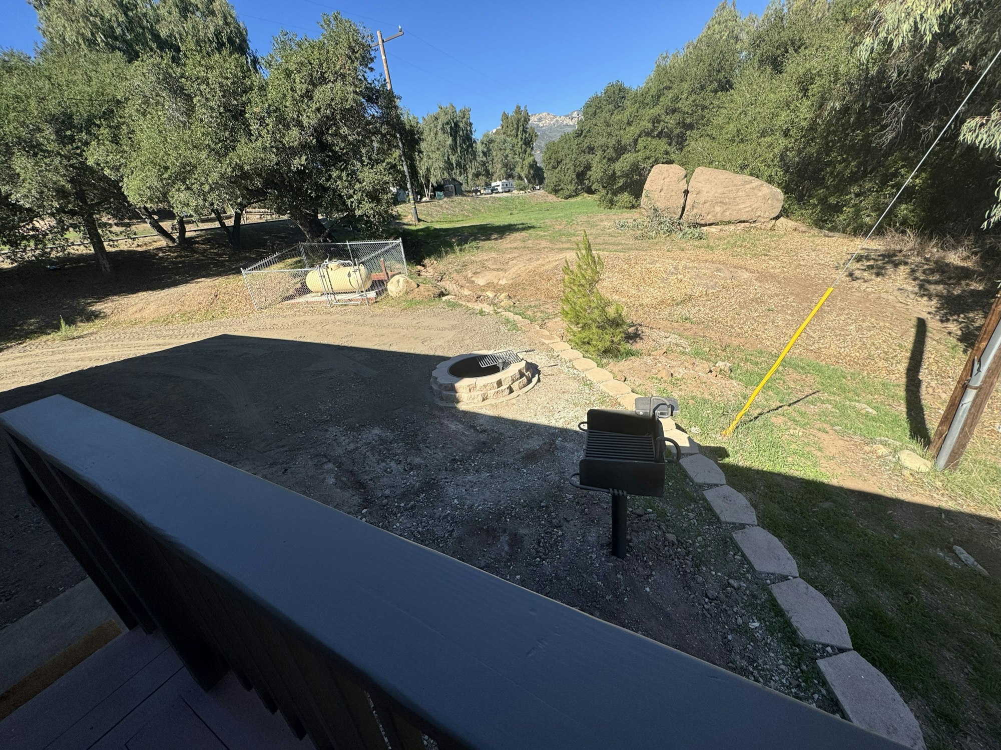 A yard with a fire pit, BBQ grill, propane tank, trees, and rocks under a clear blue sky.