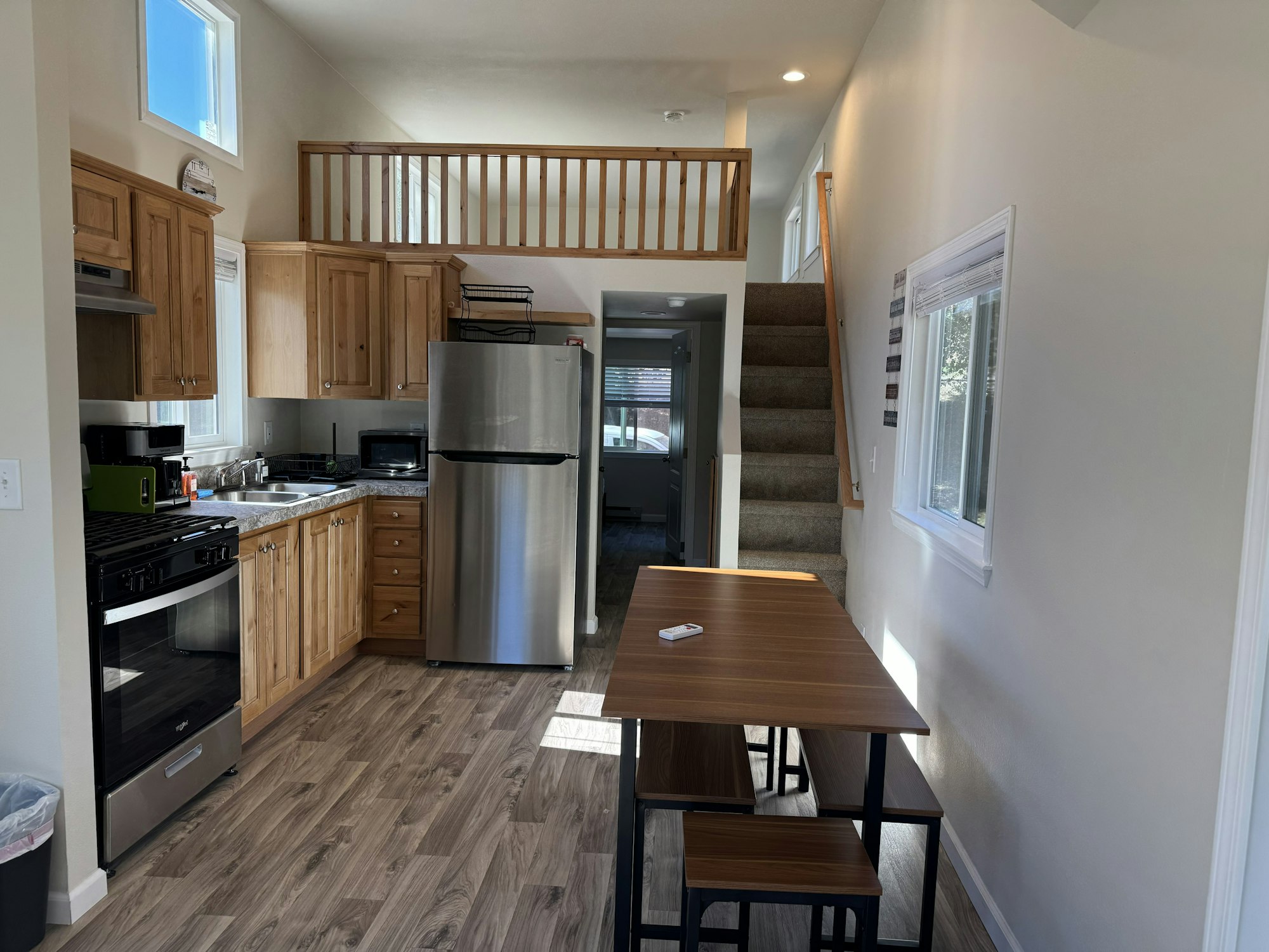 Compact kitchen with wooden cabinets, stainless steel appliances, small dining table, stairs, and loft area.