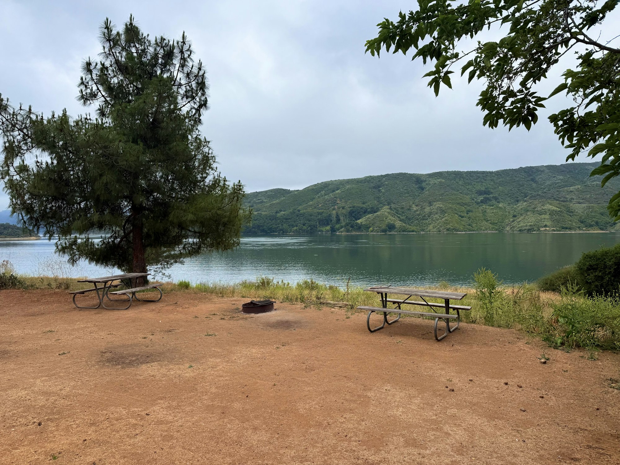 A lakeside campsite with two picnic tables and a fire pit on reddish dirt. A large tree provides partial shade near the edge of the water, with rolling green hills visible across the lake under a cloudy sky.