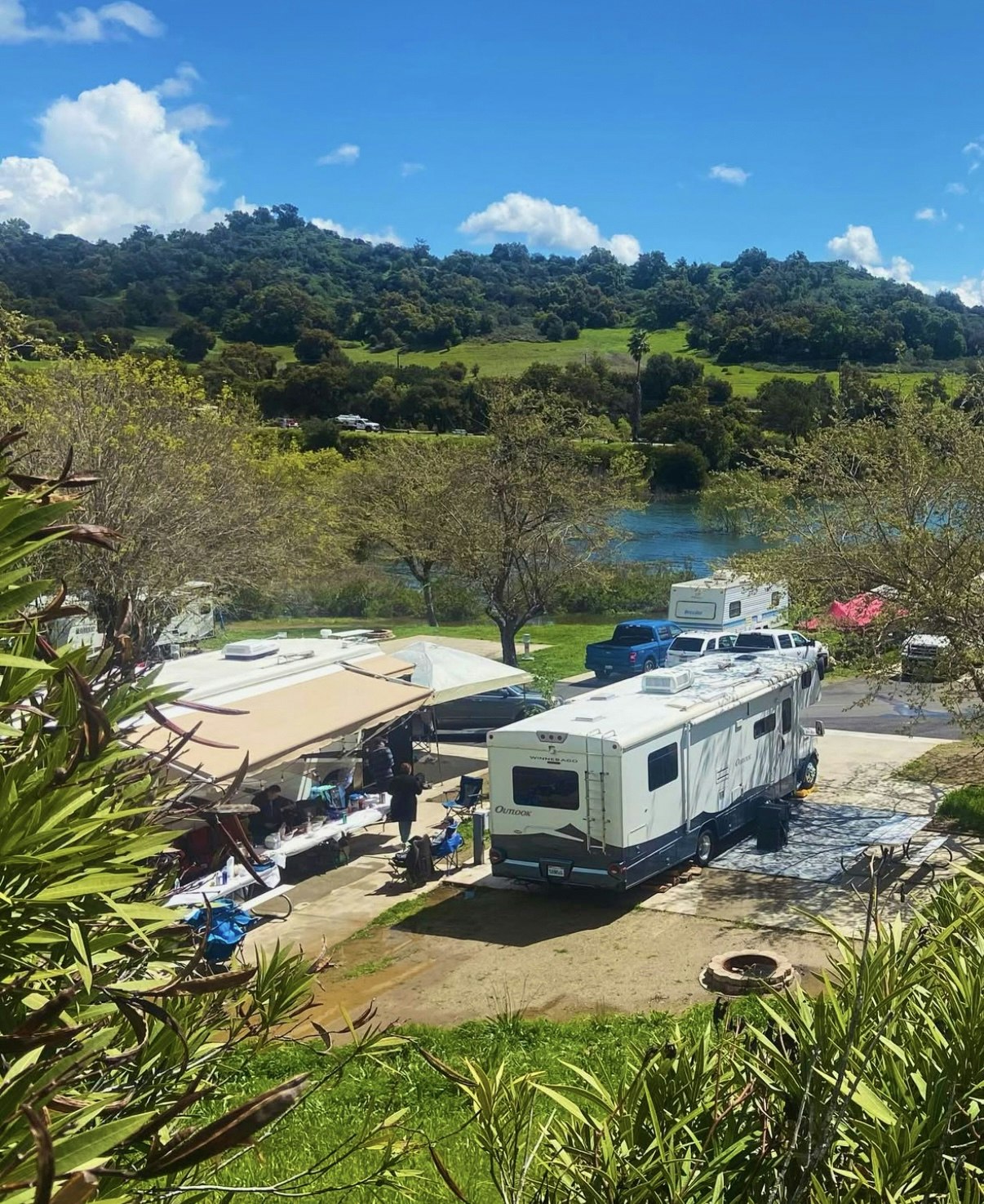 RV parked in a campground with canopies and tables, near a river and green hillside under a blue sky.