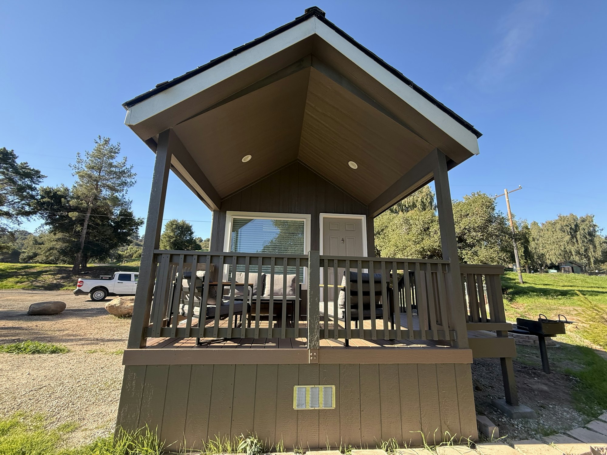 A small cabin with a porch, surrounded by greenery and a gravel path, featuring outdoor seating and a grill nearby.