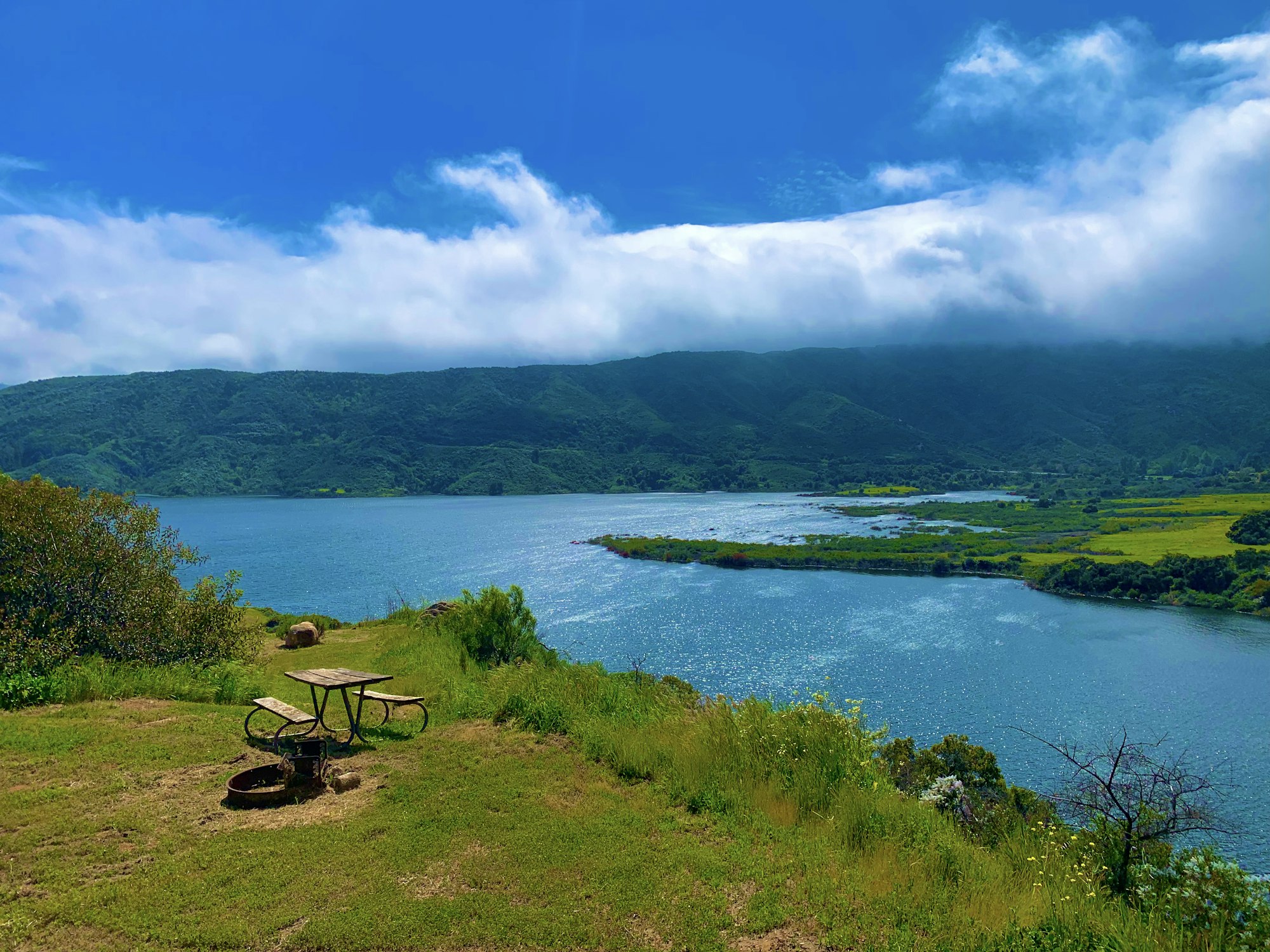 A scenic view of a lake with surrounding green hills, a cloudy sky, and a picnic table with a fire pit in the foreground.