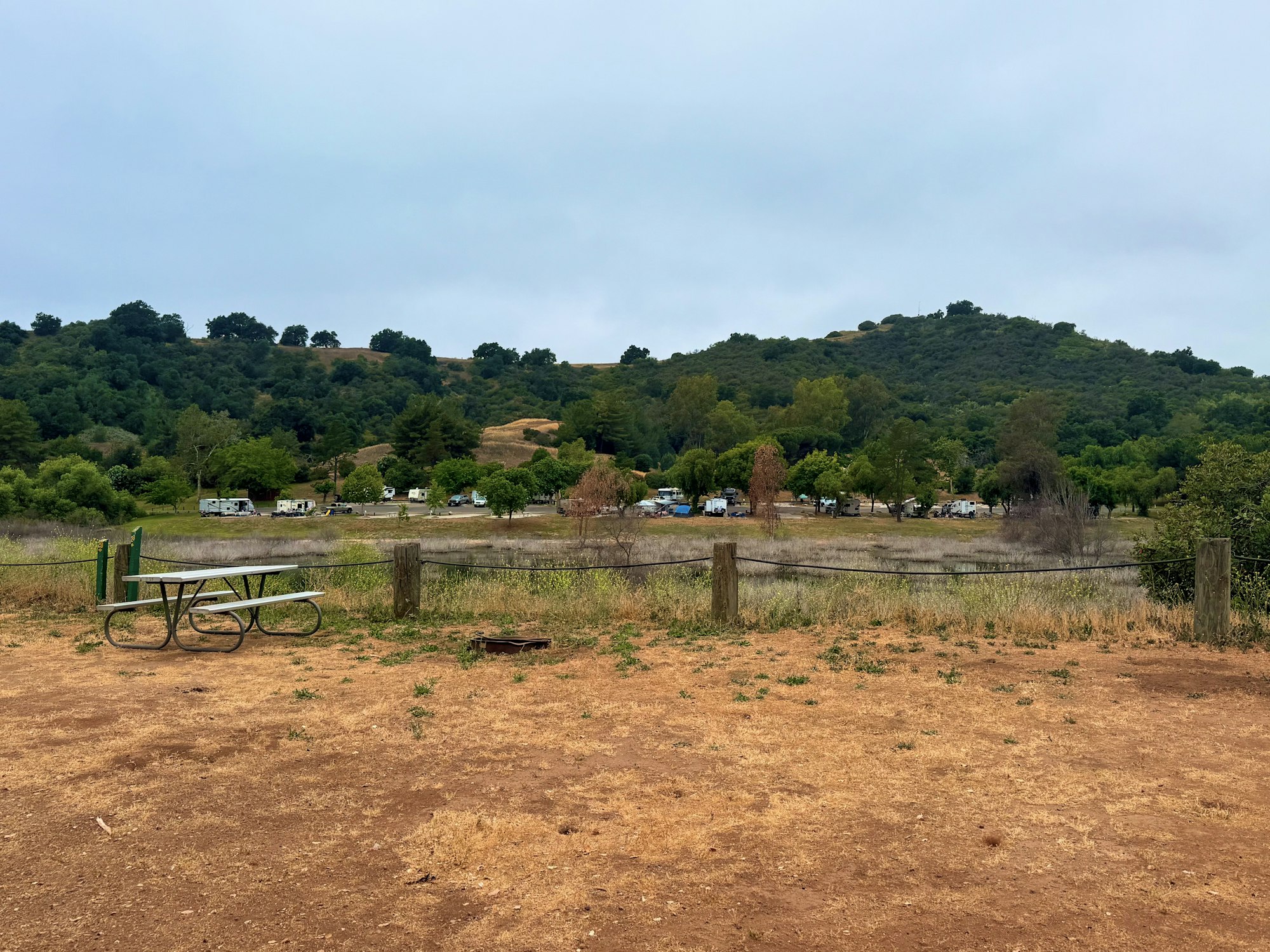 A dry, open campsite with a green picnic table and fire ring on reddish-brown dirt. In the background, there is a small body of water and a row of cabins or trailers at the base of a green hillside under a cloudy sky.