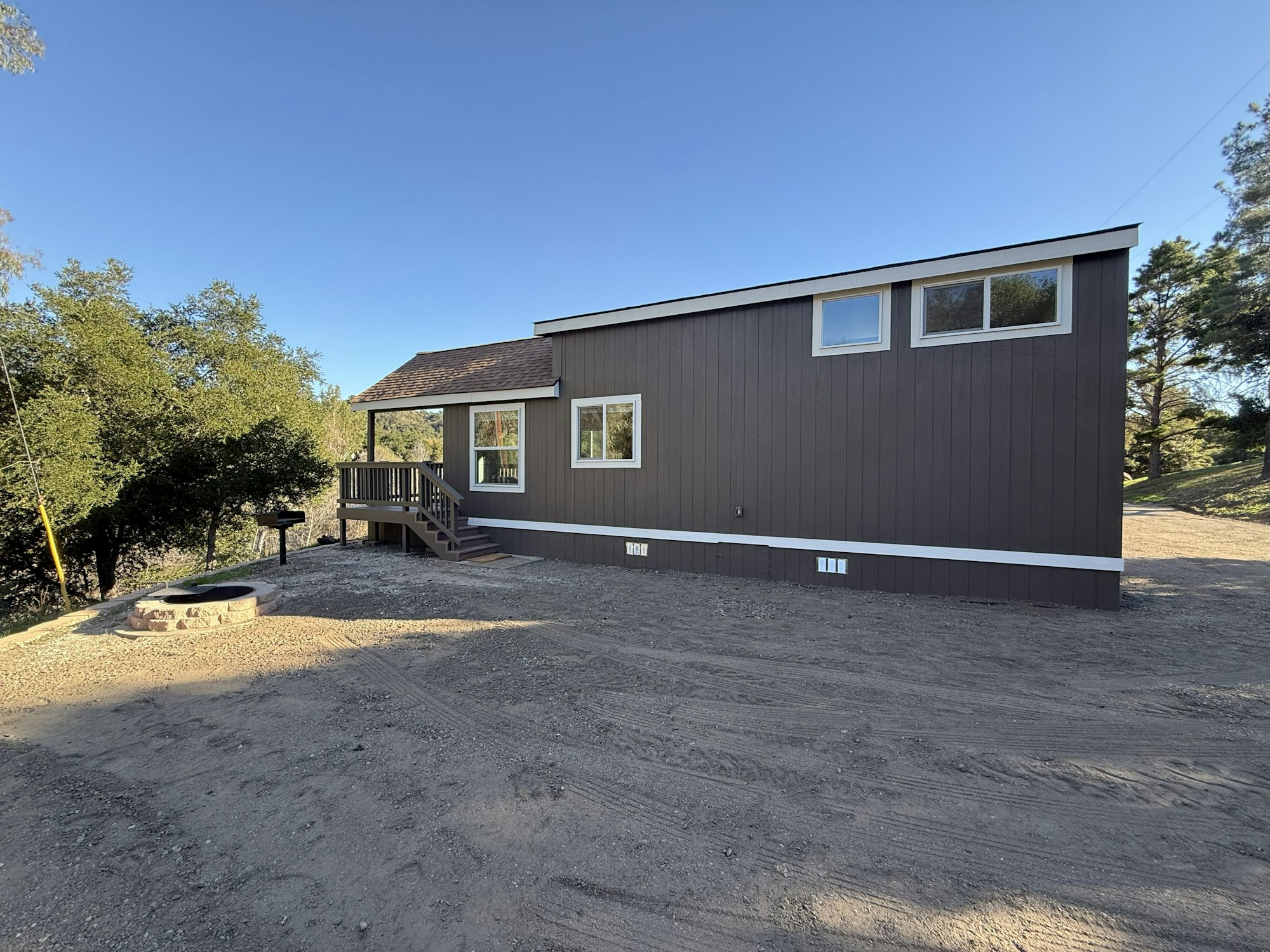 A small brown house with white trim, surrounded by trees, a gravel area, and a fire pit.