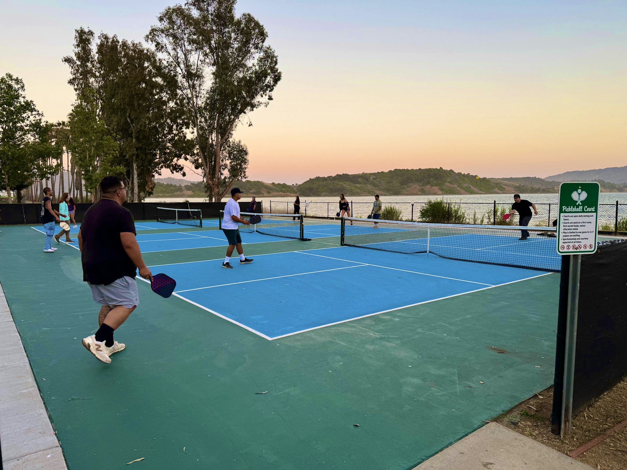 People playing pickleball on a court near a lake at sunset.
