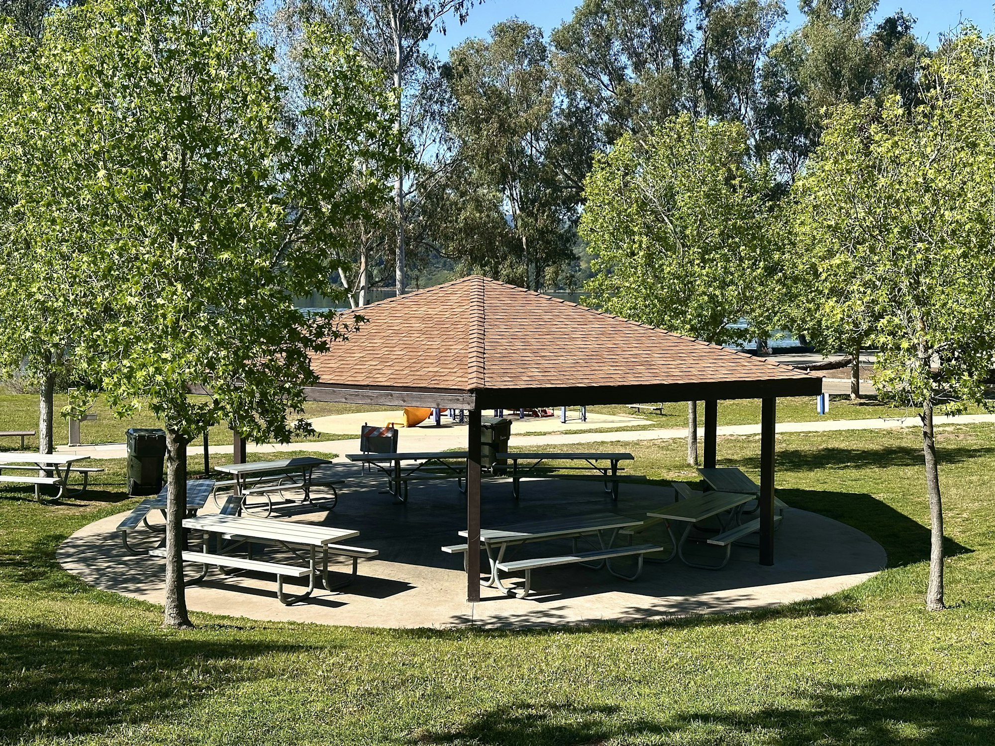A park pavilion with picnic tables surrounded by trees and grass on a sunny day.