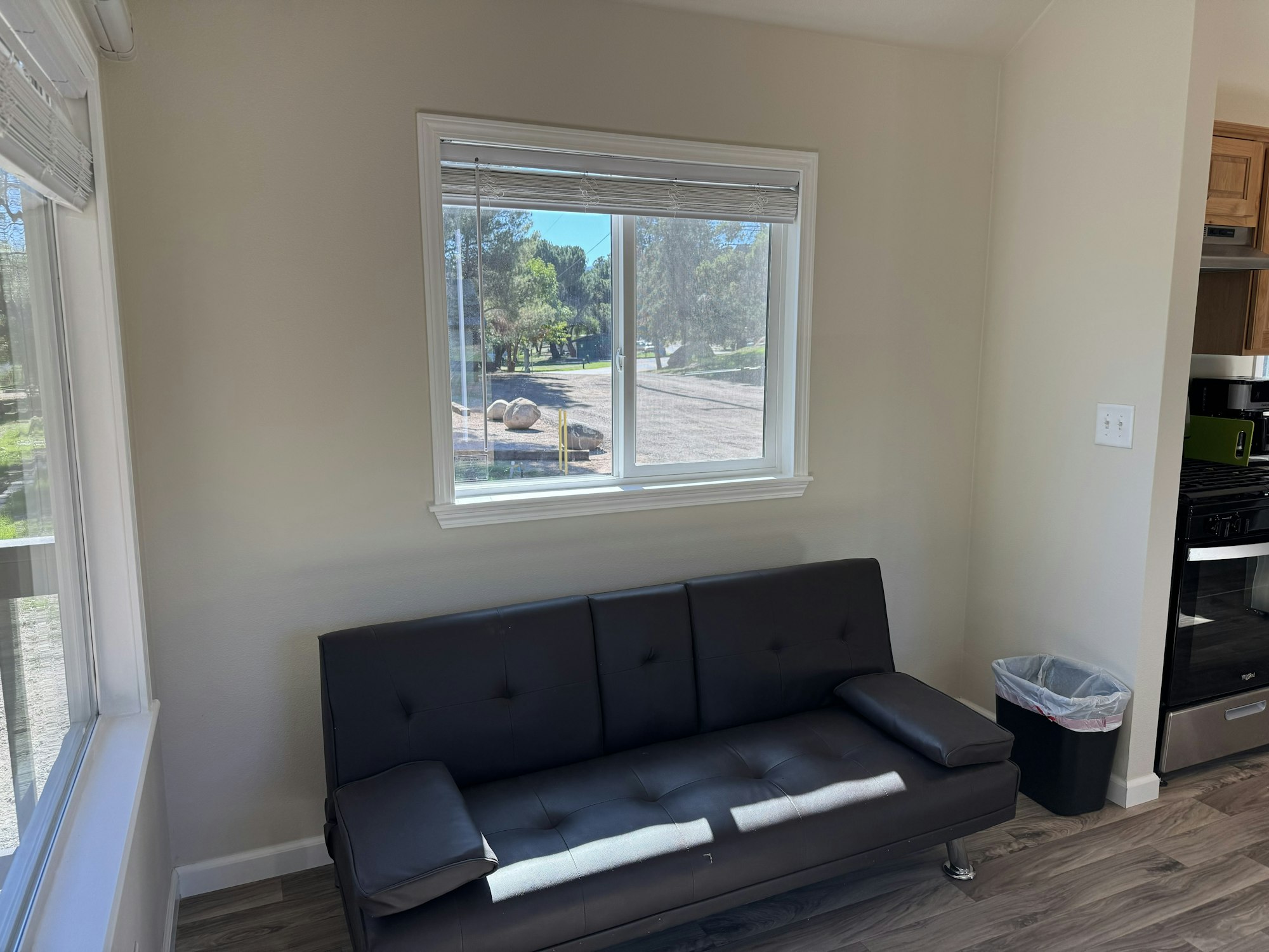 A living room with a black sofa, large windows showing a scenic view, a trash bin, and part of a kitchen area.