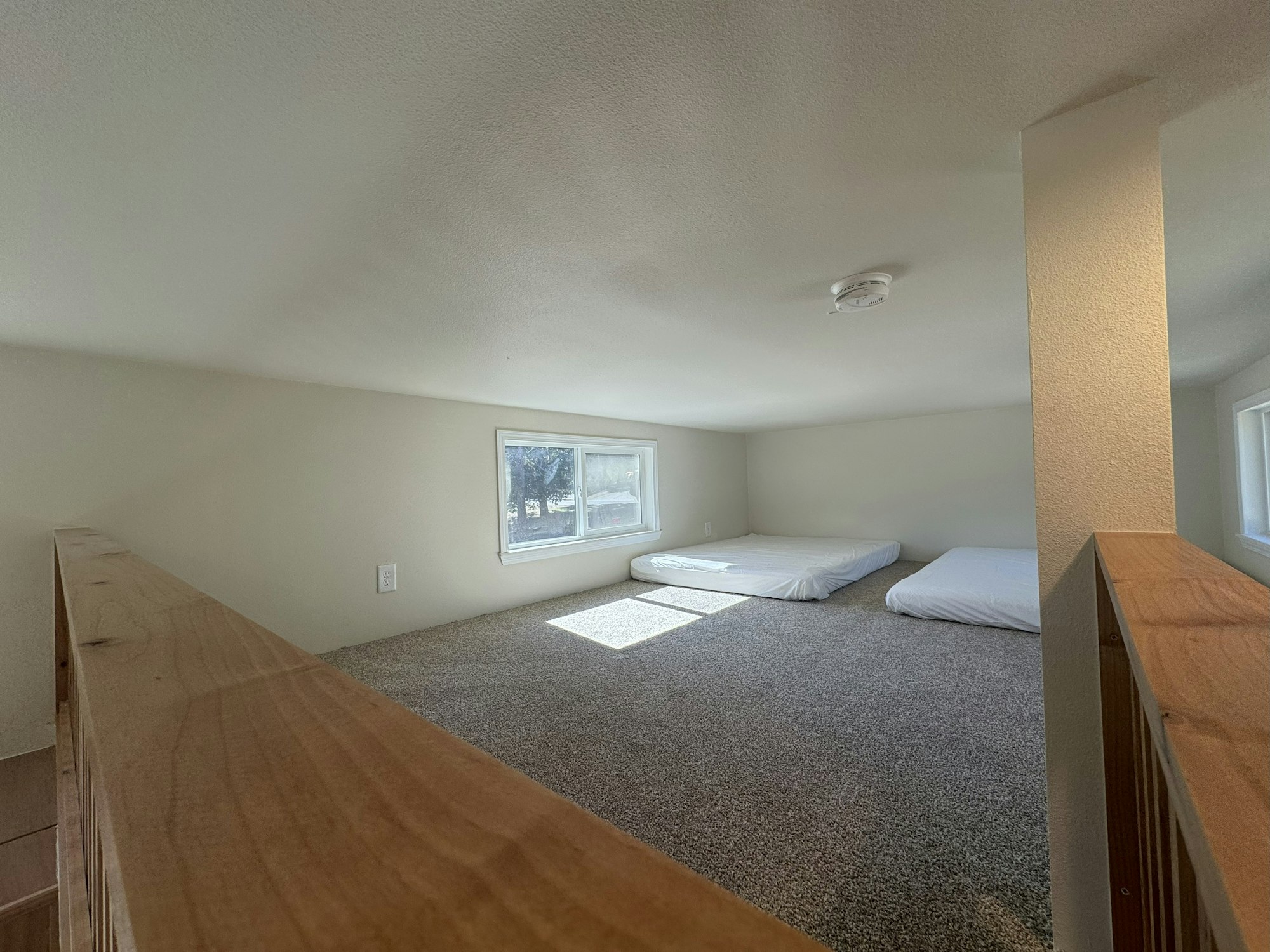 A small loft with low ceiling, carpeted floor, two mattresses, and windows letting in natural light.