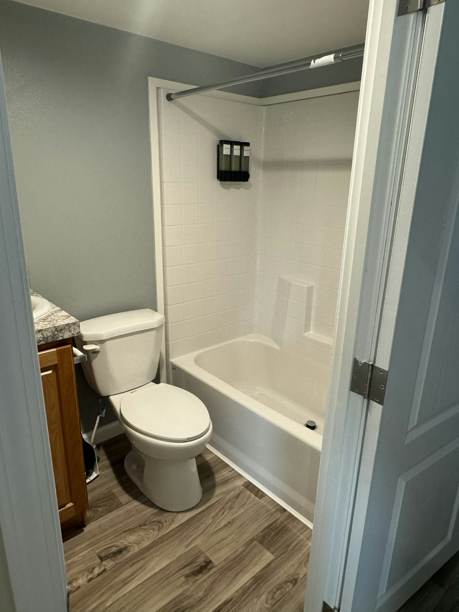 Small bathroom with toilet, bathtub, wall-mounted dispensers, and wood-style flooring.