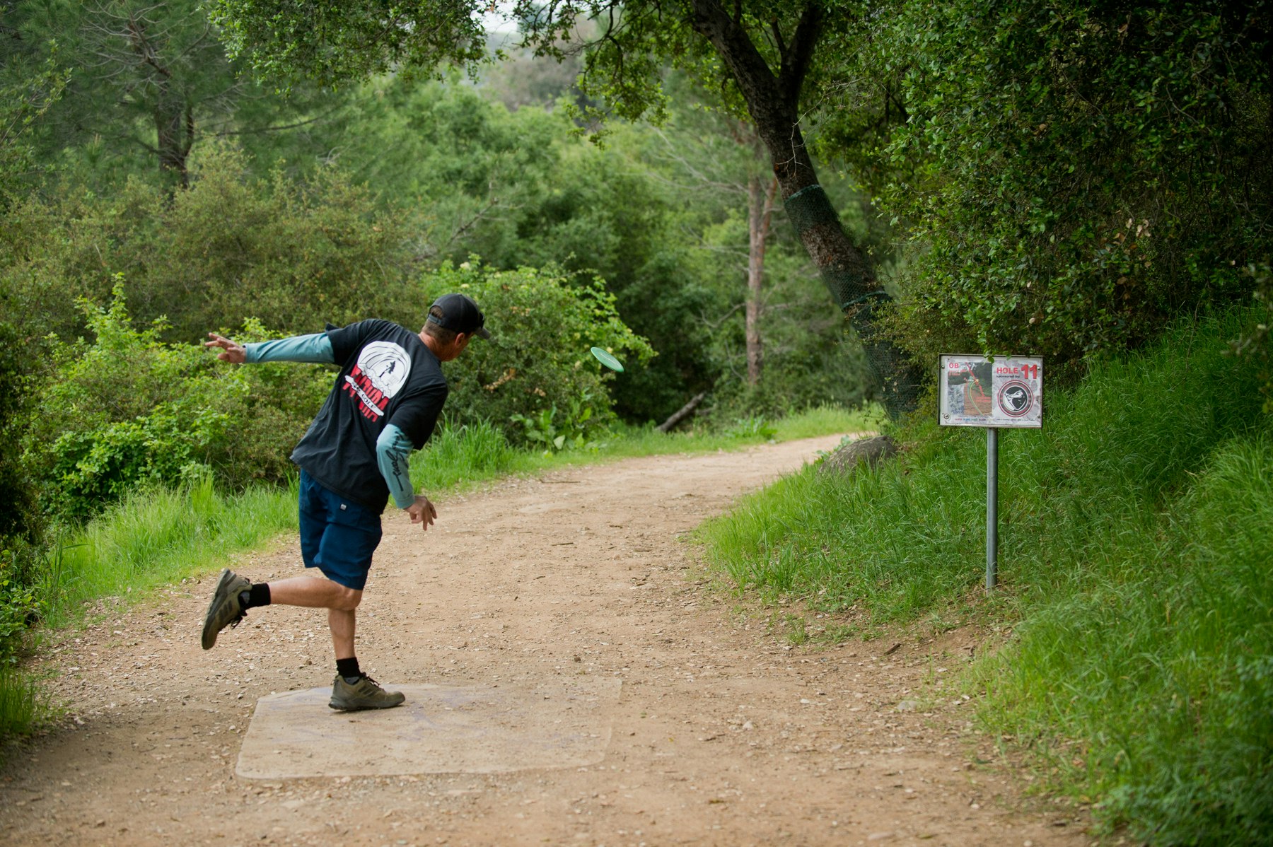 A person throwing a disc on a wooded path near a hole 11 disc golf sign.