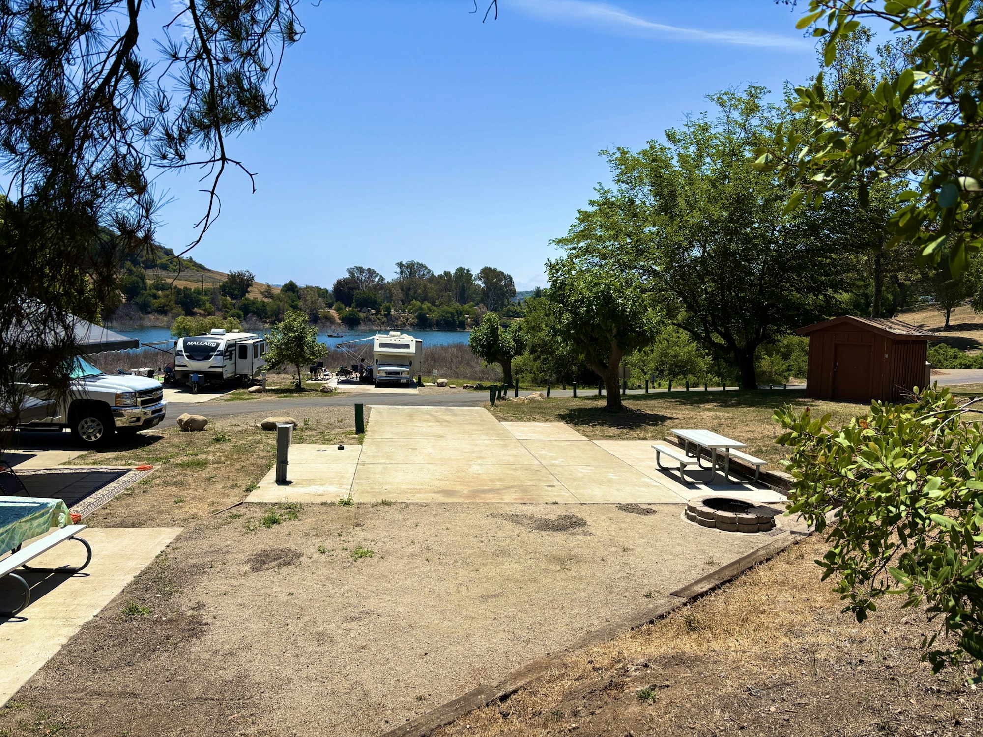 Campsite with picnic table, fire pit, RVs, truck, trees, and a lake view in the background.