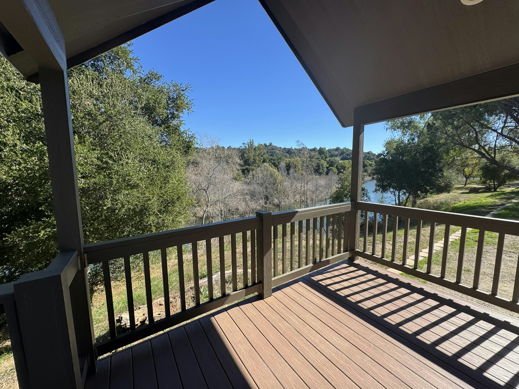 A wooden porch overlooks a scenic view with trees, a path, and a body of water under a clear blue sky.