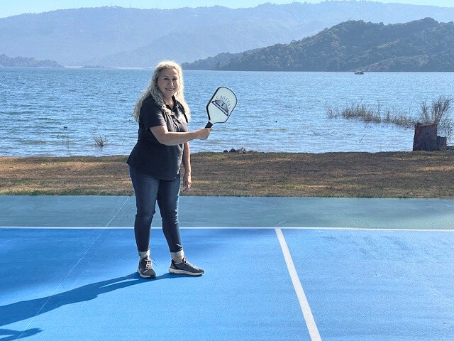 A woman with a paddle on a blue court by a lake.