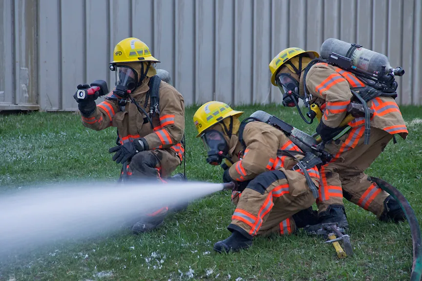 Three firefighters in protective gear are actively using hoses to spray water, likely training or responding to a fire situation.