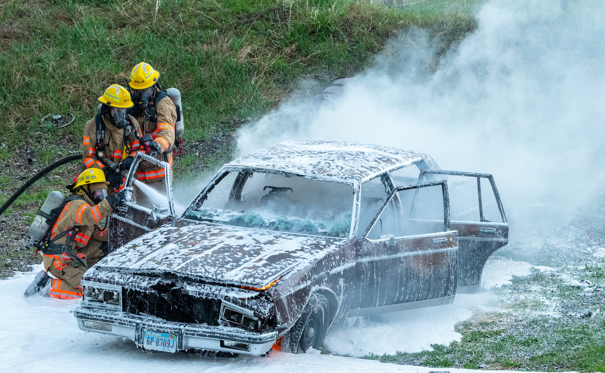 Firefighters extinguishing a vehicle fire using foam, with smoke and fire apparatus visible.