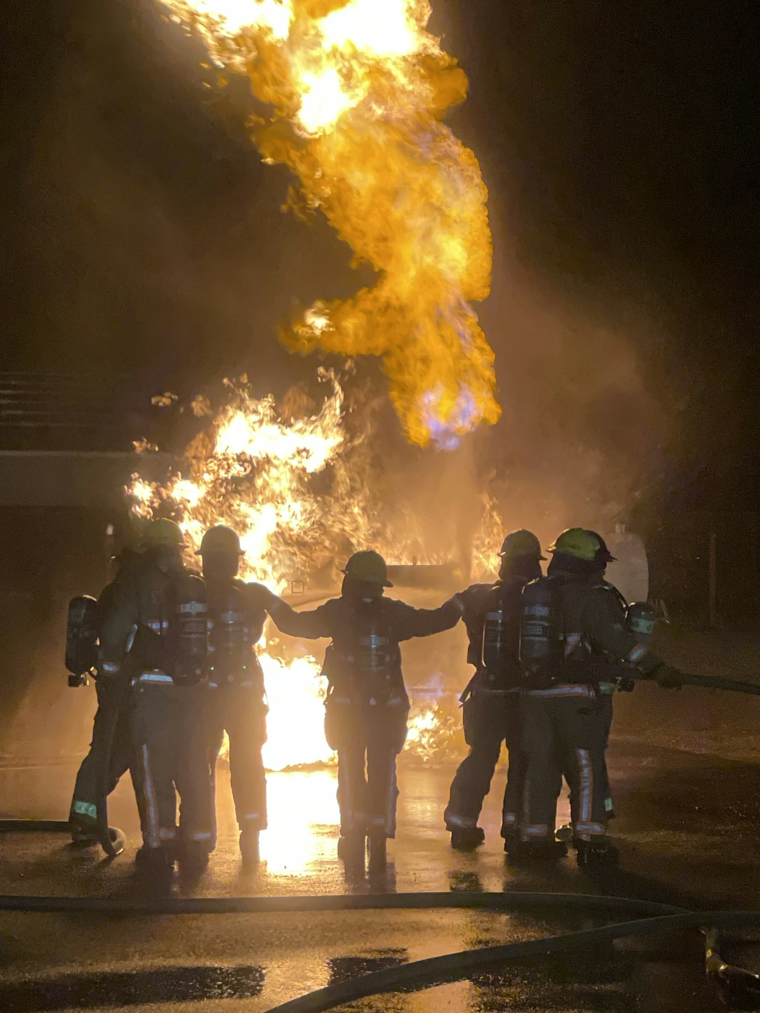 Firefighters in protective gear stand against a backdrop of flames, actively responding to a fire. Intense and dramatic scene.