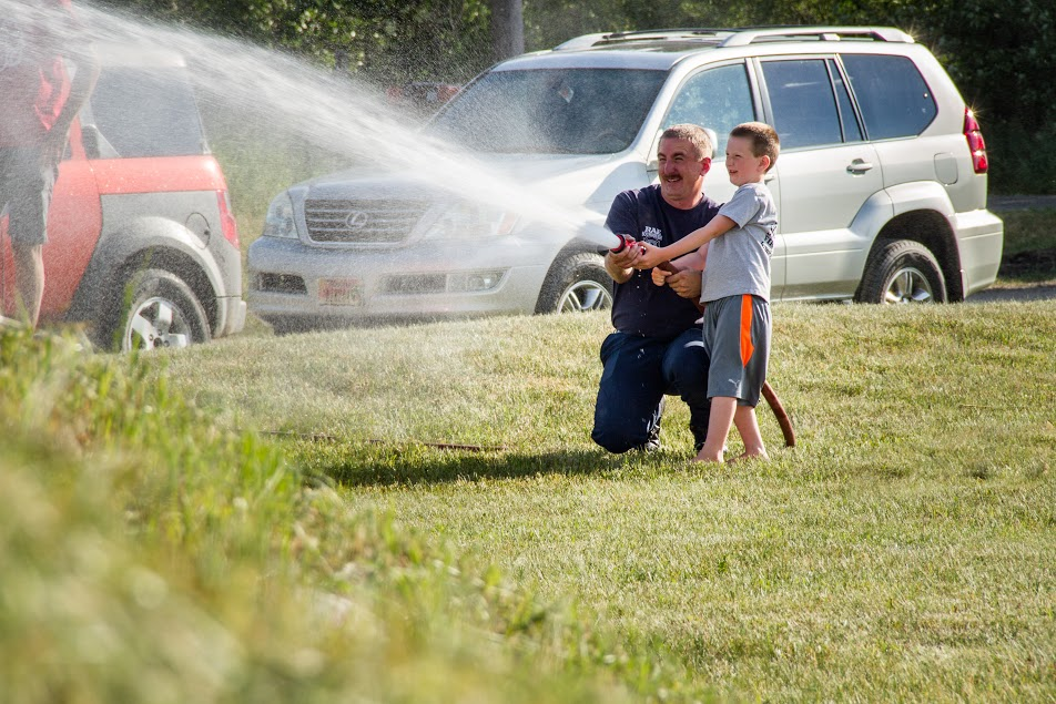 A man and a boy joyfully spray water from a hose in a grassy area, enjoying a fun moment together.