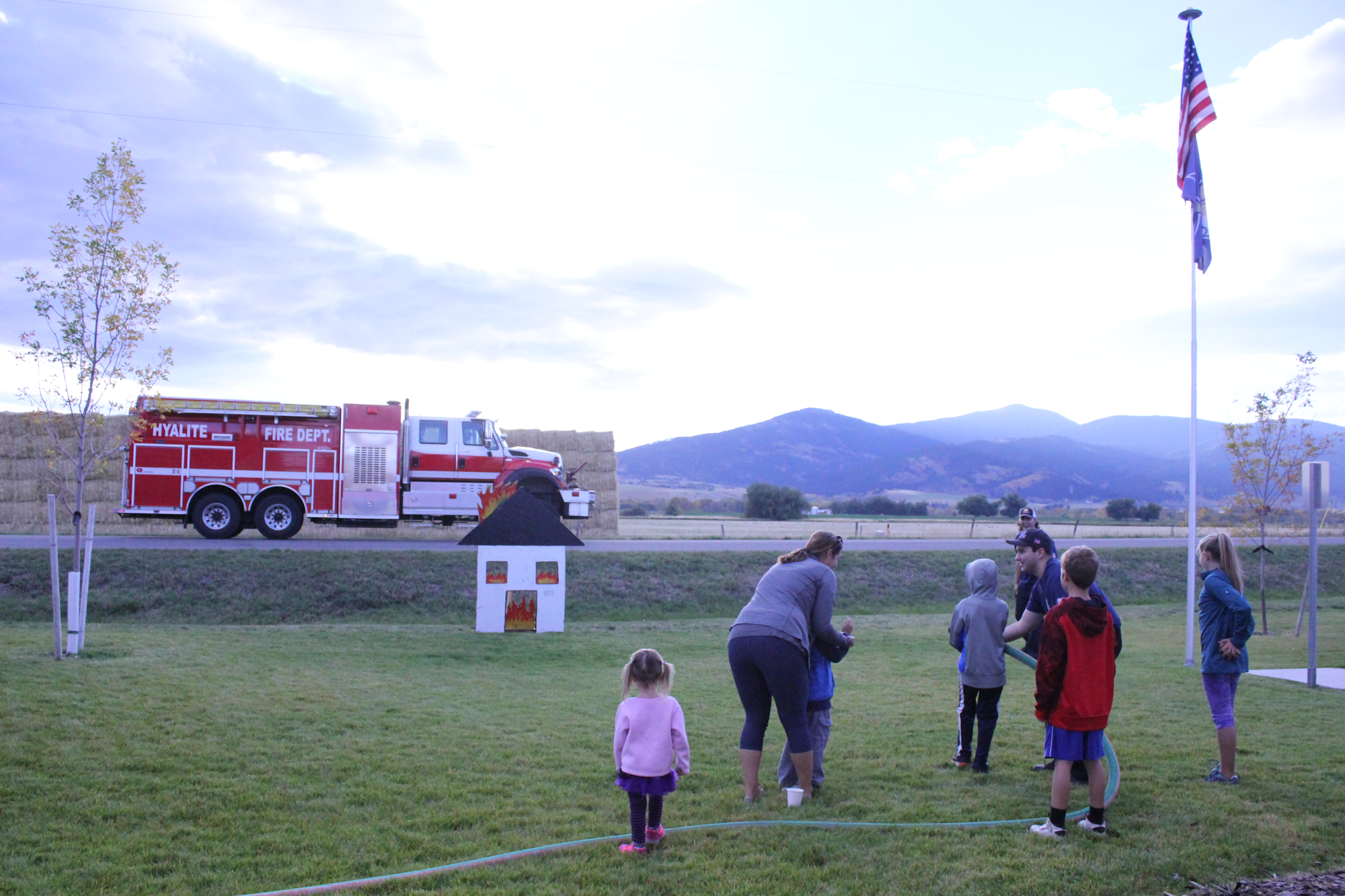 A fire truck is parked nearby while families and children engage in activities on a grassy area with mountains in the background.