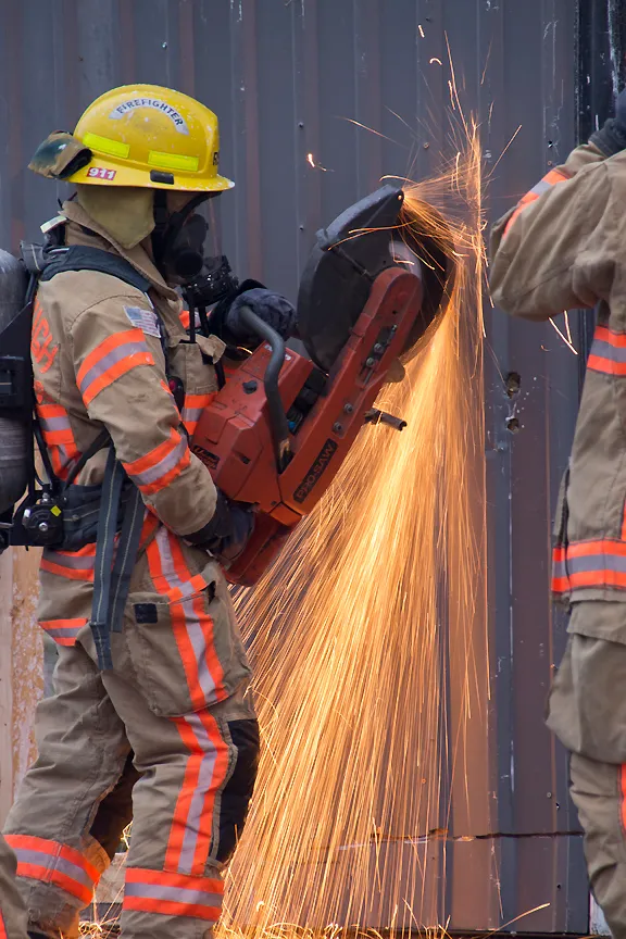 A firefighter cuts through metal with a power tool, creating sparks, while another firefighter assists nearby.