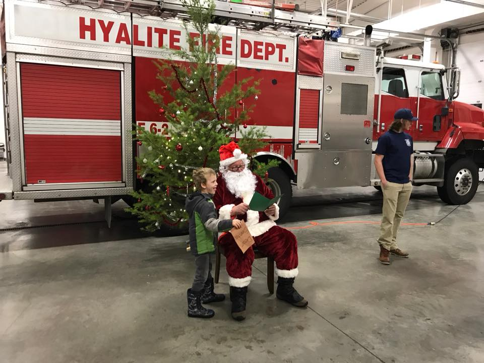 A child is meeting Santa Claus in front of a fire truck, with a decorated Christmas tree nearby.