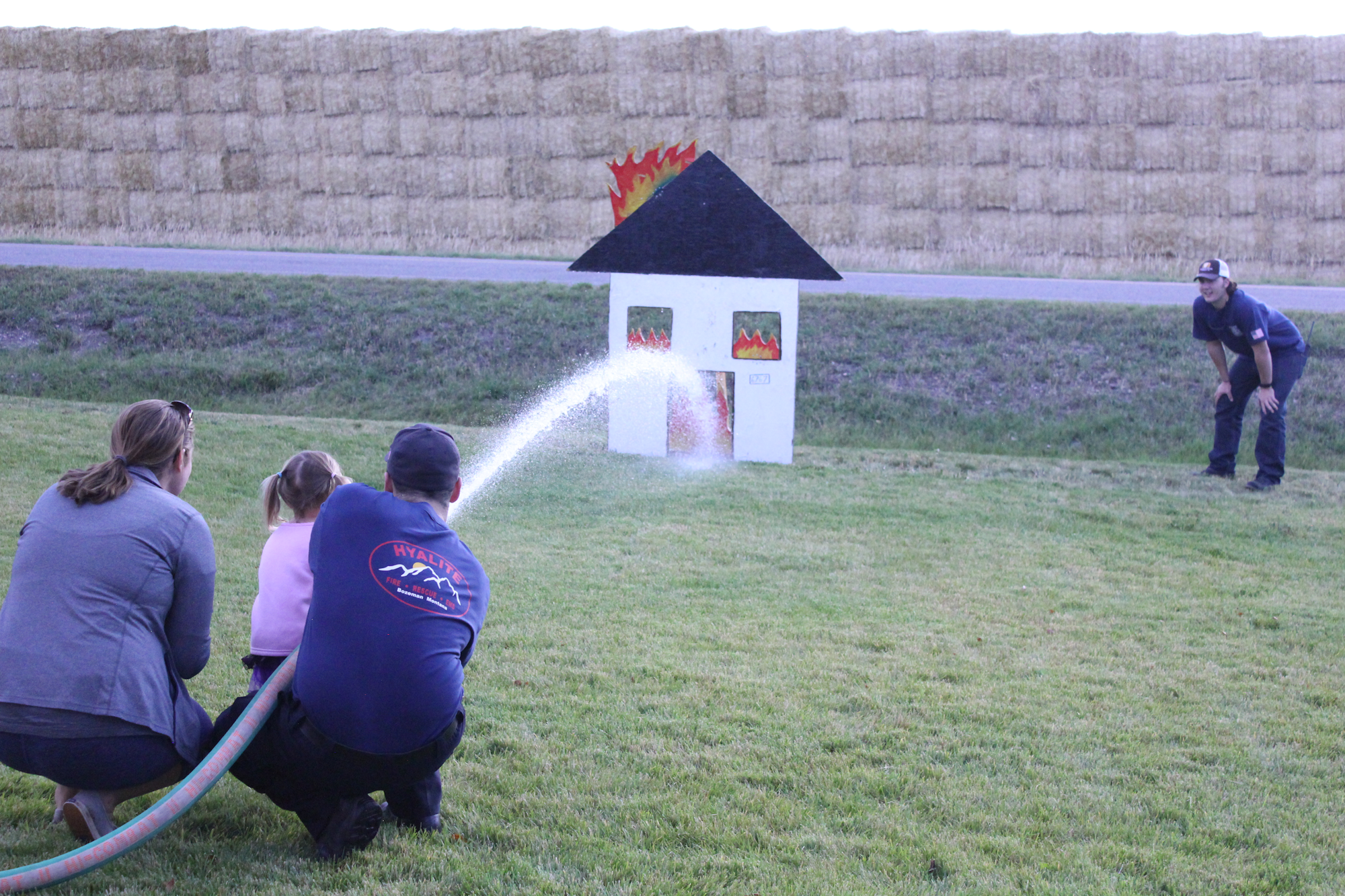 A family practices firefighting skills, spraying water at a mock house with flames, while a firefighter supervises.