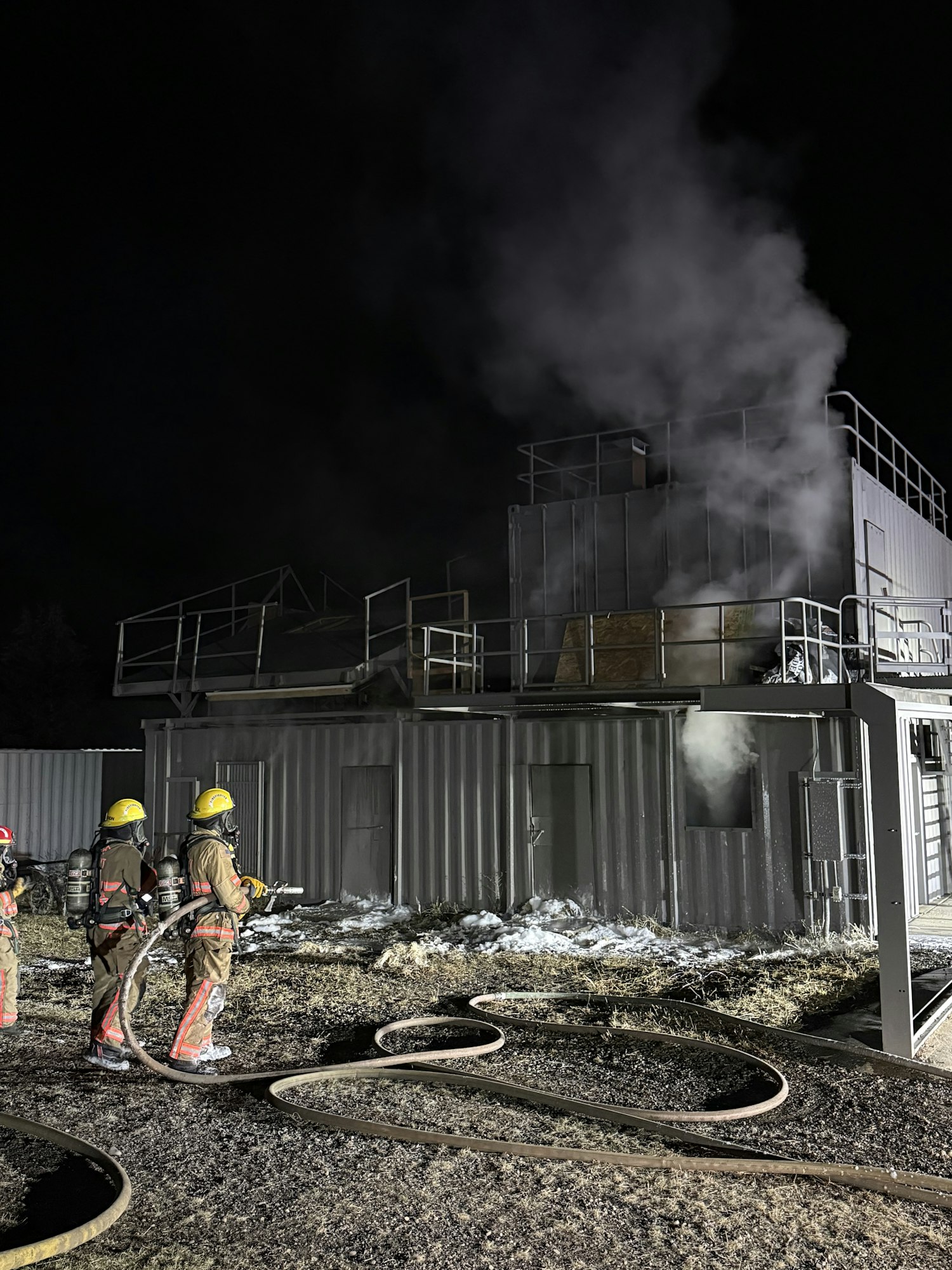 Firefighters are responding to smoke from a building at night, with hoses and equipment visible on the ground.