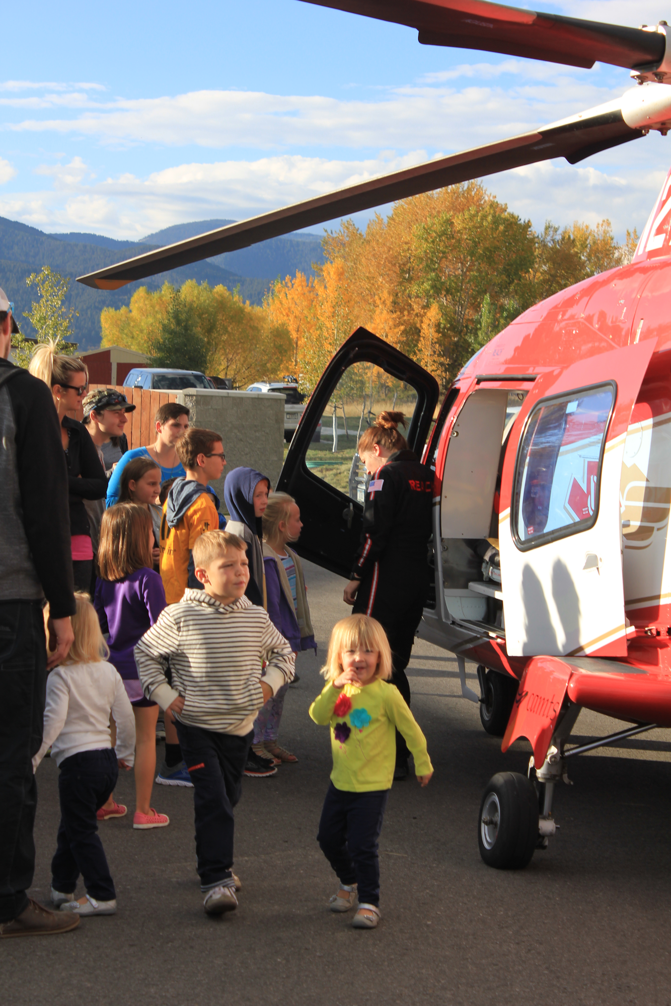 A group of children and adults gather around a helicopter, with a crew member preparing for an event or demonstration.