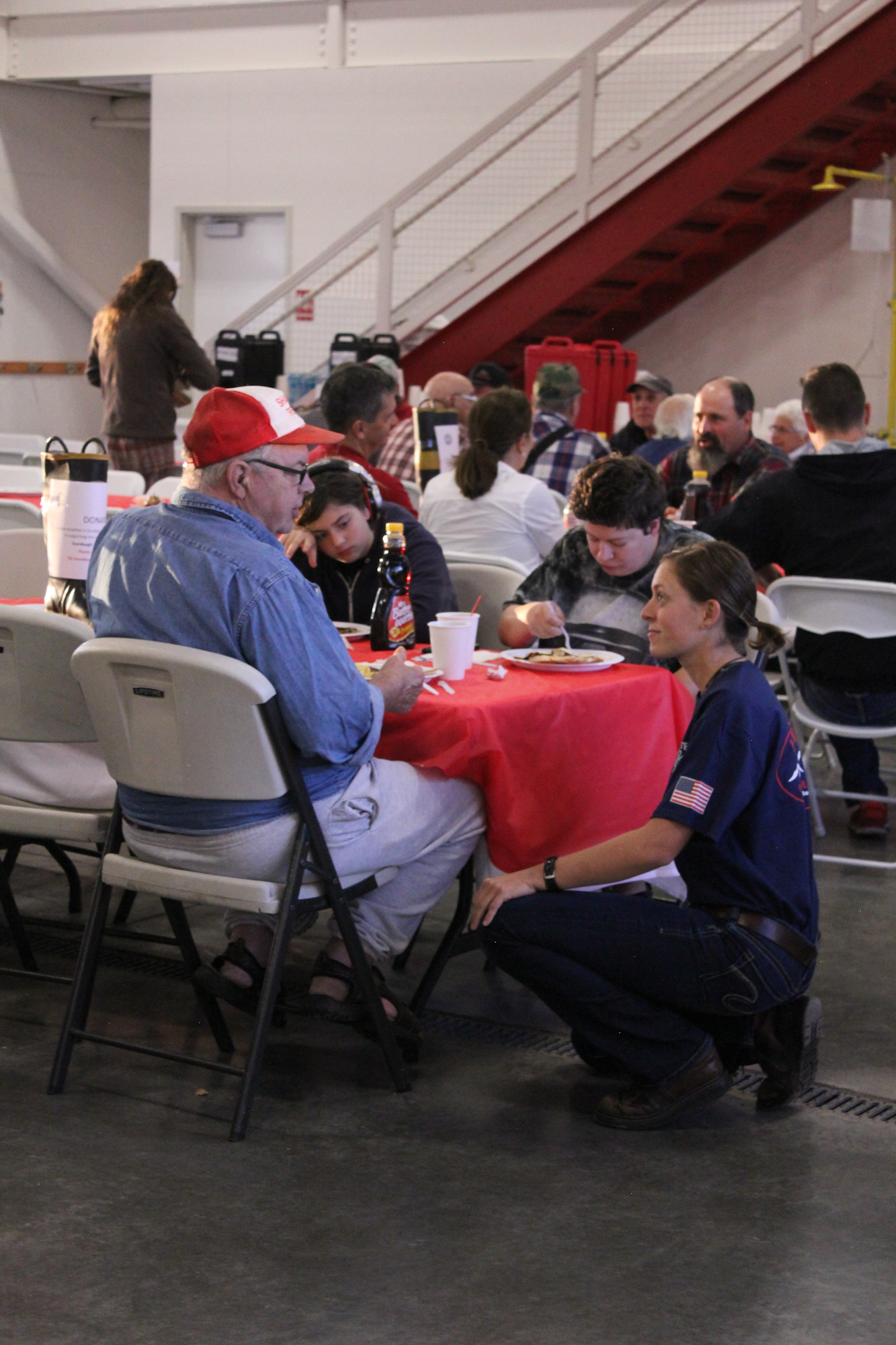 A group of people at a communal table sharing a meal, with one woman kneeling to speak with an older man and a child.