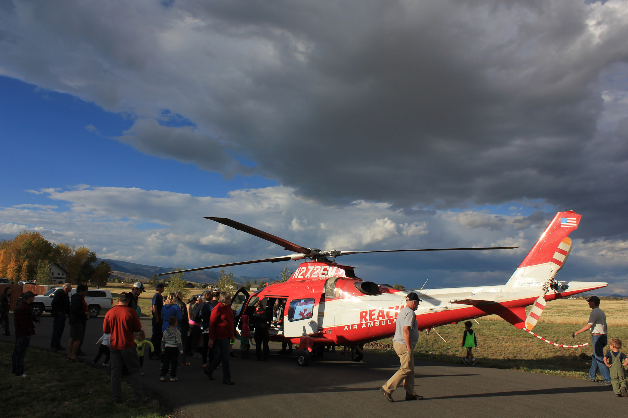 A crowd gathers around a red and white air ambulance helicopter on a cloudy day, with people interacting and observing.